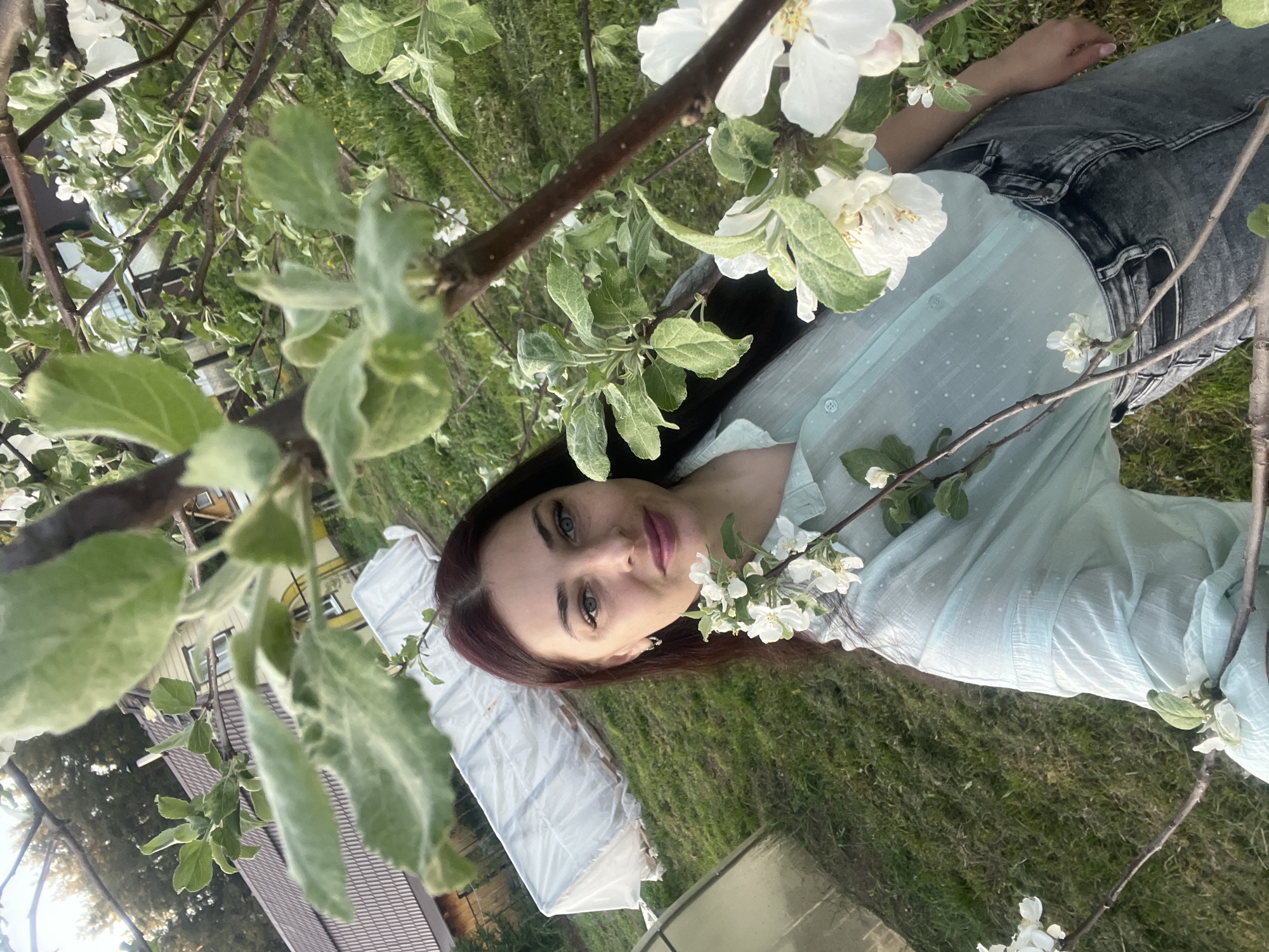 The image shows a woman lying on a grassy area. Her hair is dark red. She is wearing a light blue shirt and gray jeans. She is surrounded by branches with white flowers and green leaves. A white sheet is visible behind her to the left. A wooden building is partially visible in the background. The location is Garevaya, Russia.