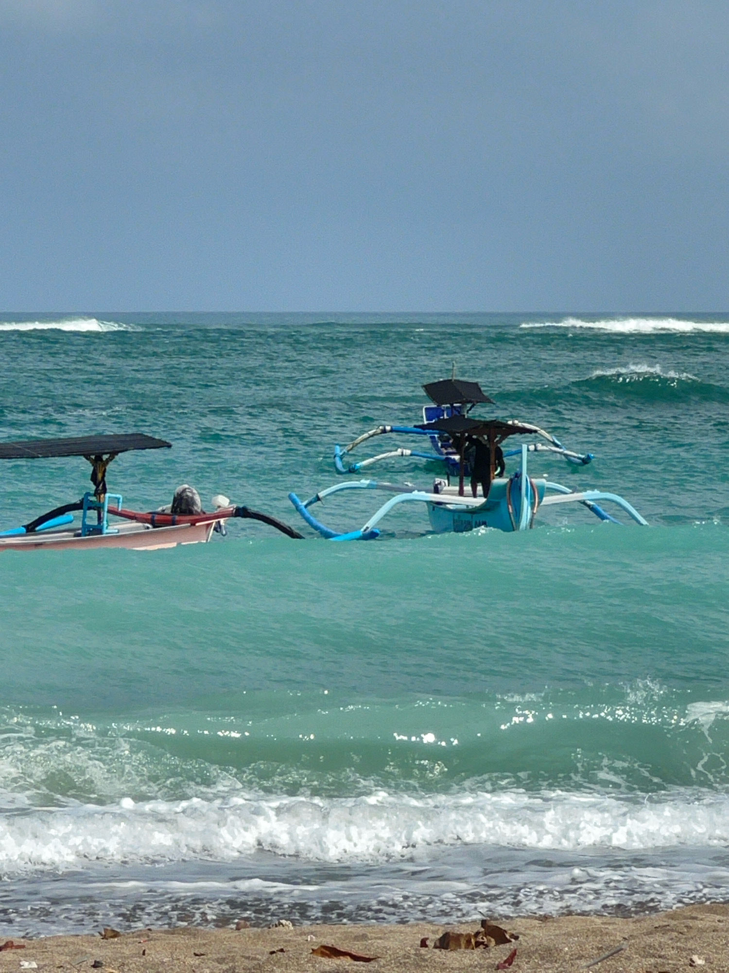 The image shows a bright day at the beach with two traditional Balinese outrigger boats in the turquoise water off the coast of Kuta, Indonesia. The boat on the left is smaller and has a black canopy. The boat on the right is larger and also has a black canopy, with two figures visible inside. The water is choppy with waves breaking near the shore. The beach in the foreground is sandy and appears to have some debris. The sky is a pale blue.