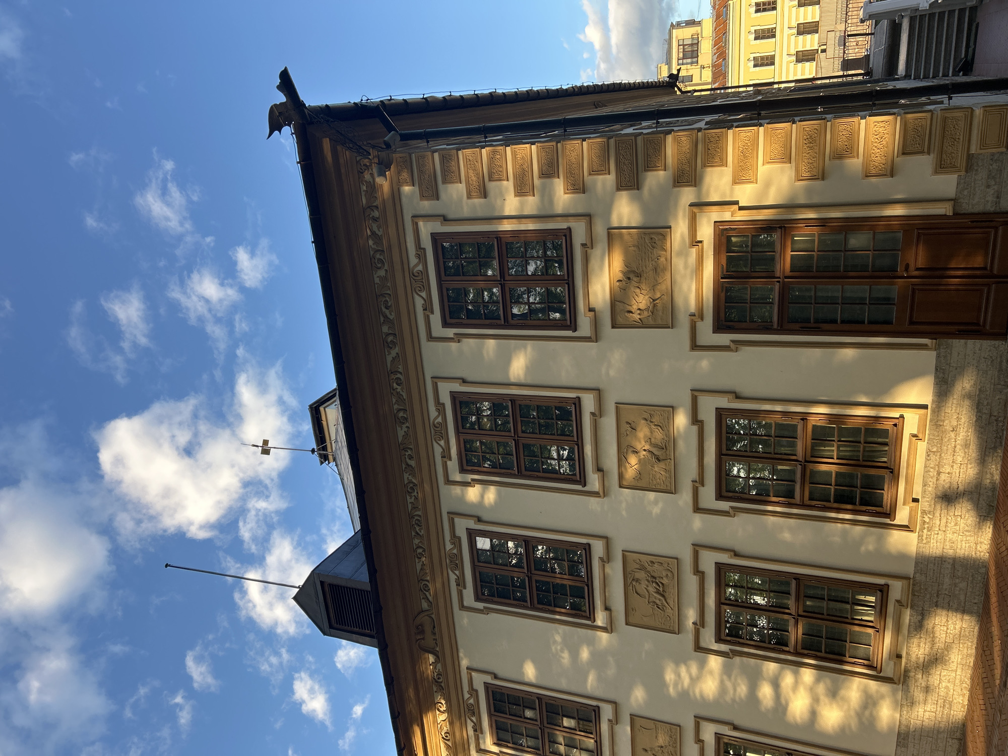 A close-up, low-angle shot captures the facade of an ornate building with a light yellow stucco exterior. The building features several tall, narrow windows with dark wooden frames and multiple panes, reflecting the surrounding environment and a bright blue sky dotted with white clouds. Between the windows, relief carvings depict scenes with figures, adding to the architectural detail. Decorative molding, also in a golden hue, frames the windows and runs along the top of the facade, creating a sense of grandeur. The building's corner is accented with a series of rectangular, embossed panels. A dark, ornate gutter spout shaped like a mythical creature protrudes from the roofline. The sun casts long, dramatic shadows across the facade, suggesting it is either early morning or late afternoon. In the background, another building with a yellow facade and visible windows can be seen, partially obscured by the foreground structure. The overall scene conveys a sense of historical architecture and detailed craftsmanship under a dynamic sky.