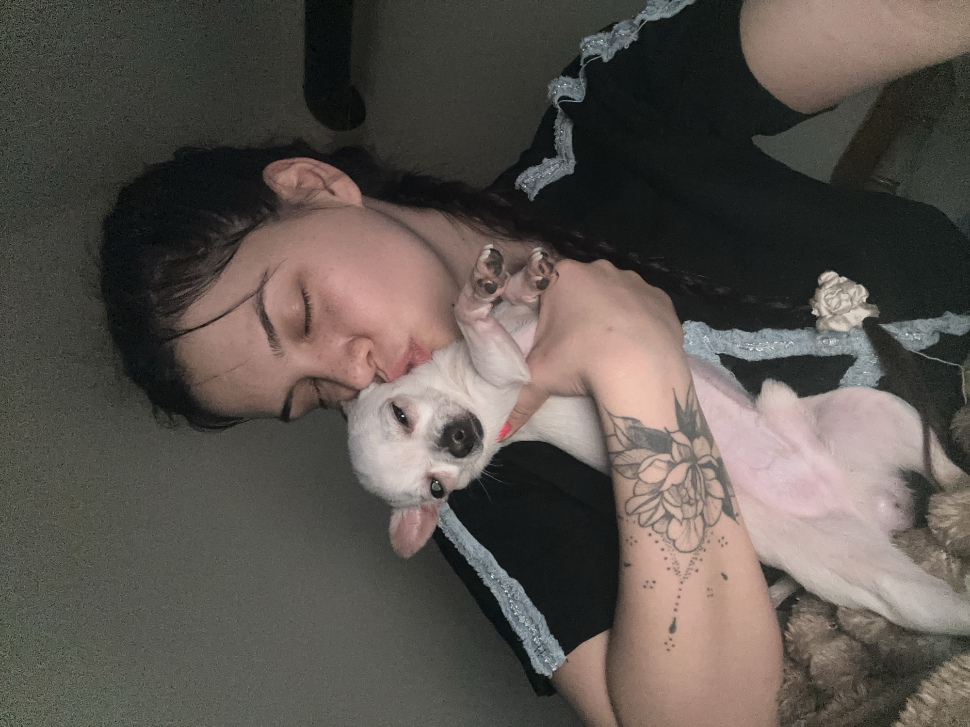 An indoor scene depicts an individual, a young adult female, holding a small white dog. The female has dark hair, styled partially in a visible braid on the right side of her head, and freckles on her nose and cheeks. Her eyes are closed or squinted, and her lips are pursed in a kissing gesture directed towards the dog's head. She wears a dark-colored garment with light blue or silver textured trim along the shoulders. A prominent floral tattoo with dotted line details is visible on her left forearm. Her right hand, with pink nail polish on one digit, supports the dog. The dog is a small, light-colored breed, predominantly white with visible pink skin on its belly and legs, and has partially open eyes looking towards the viewer. Dark-colored nails are visible on its paws. The background is a plain, dark gray wall. A textured, brown, possibly fabric, object is partially visible in the lower left corner, while a dark, cylindrical object is partially visible on the far right. The scene is located in the city of Chubra, Bulgaria.