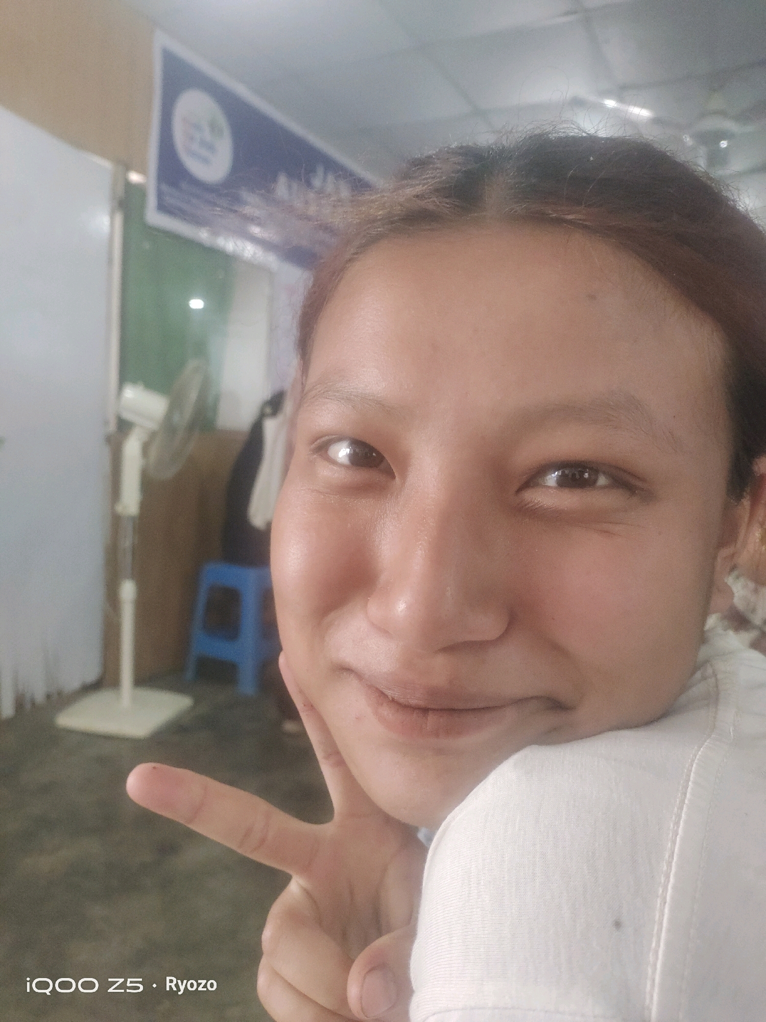 A close-up selfie captures a young woman with light brown hair, her face filling most of the frame. She is smiling gently, resting her head on her hand, and making a peace sign with her other hand. Her eyes are open and looking towards the camera. She is wearing a white top. In the background, out of focus, a white oscillating fan stands to the left, and a blue stool is visible behind it. A sign with a blue border and a circular logo is mounted on the wall above the fan. The overall lighting suggests an indoor setting, possibly a room in Kangpokpi, India. The timestamp at the bottom of the image reads "iQOO Z5 · Ryozo".