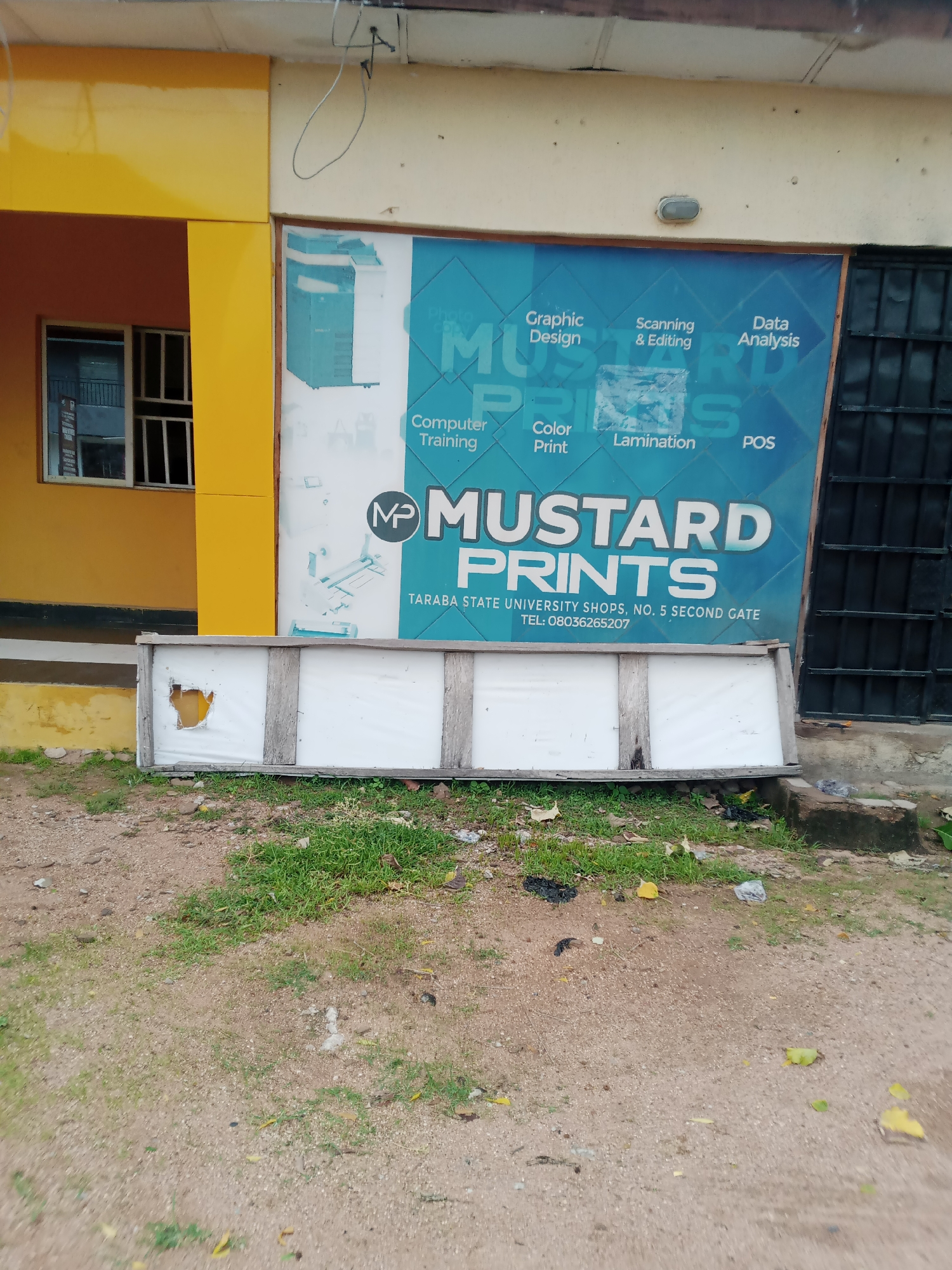 The image captures a storefront for "MUSTARD PRINTS" located in Jalingo, Nigeria, specifically at TARABA STATE UNIVERSITY SHOPS, NO. 5 SECOND GATE. The storefront features a large, blue sign with white lettering that advertises various services including "Graphic Design," "Scanning & Editing," "Data Analysis," "Computer Training," "Color Print," "Lamination," and "POS." To the left of the sign, a portion of a yellow building with a window is visible. In front of the storefront, a damaged white board with a wooden frame is leaning on the ground, partially obscuring the lower part of the sign. The ground in front of the building is a mixture of dirt and sparse green grass, with some scattered debris. A black metal gate is visible to the right of the sign. The overall scene depicts a commercial establishment in an outdoor urban environment.