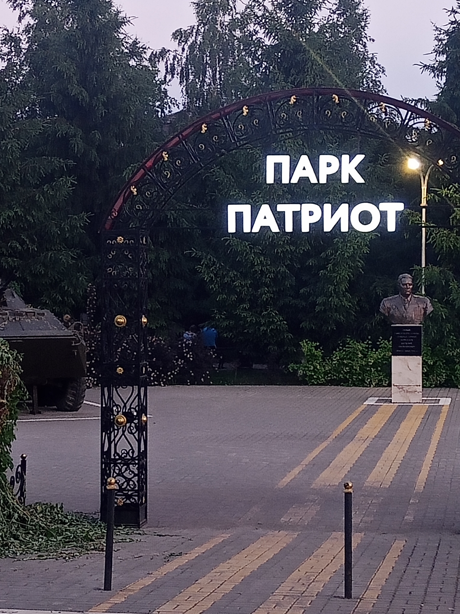The image shows an outdoor scene in front of an entrance to "PARK PATRIOT" in Panino, Russia. A large, ornate black archway with gold accents spans the entrance. White, illuminated letters on the archway spell out "ПАРК" (Park) on the top line and "ПАТРИОТ" (Patriot) on the bottom line.
To the right of the entrance, a bust of a man in military attire is displayed on a pedestal. The background is filled with dense evergreen trees. A paved pathway with yellow striped crosswalk markings leads into the park.
On the left side of the image, partially visible, is the rear of a military vehicle, possibly an armored personnel carrier. A decorative black metal gate pillar stands in the foreground on the left. A few individuals, dressed in casual clothing, are visible in the middle ground, near the trees and behind the gate. The overall lighting suggests it is either early morning or late evening.