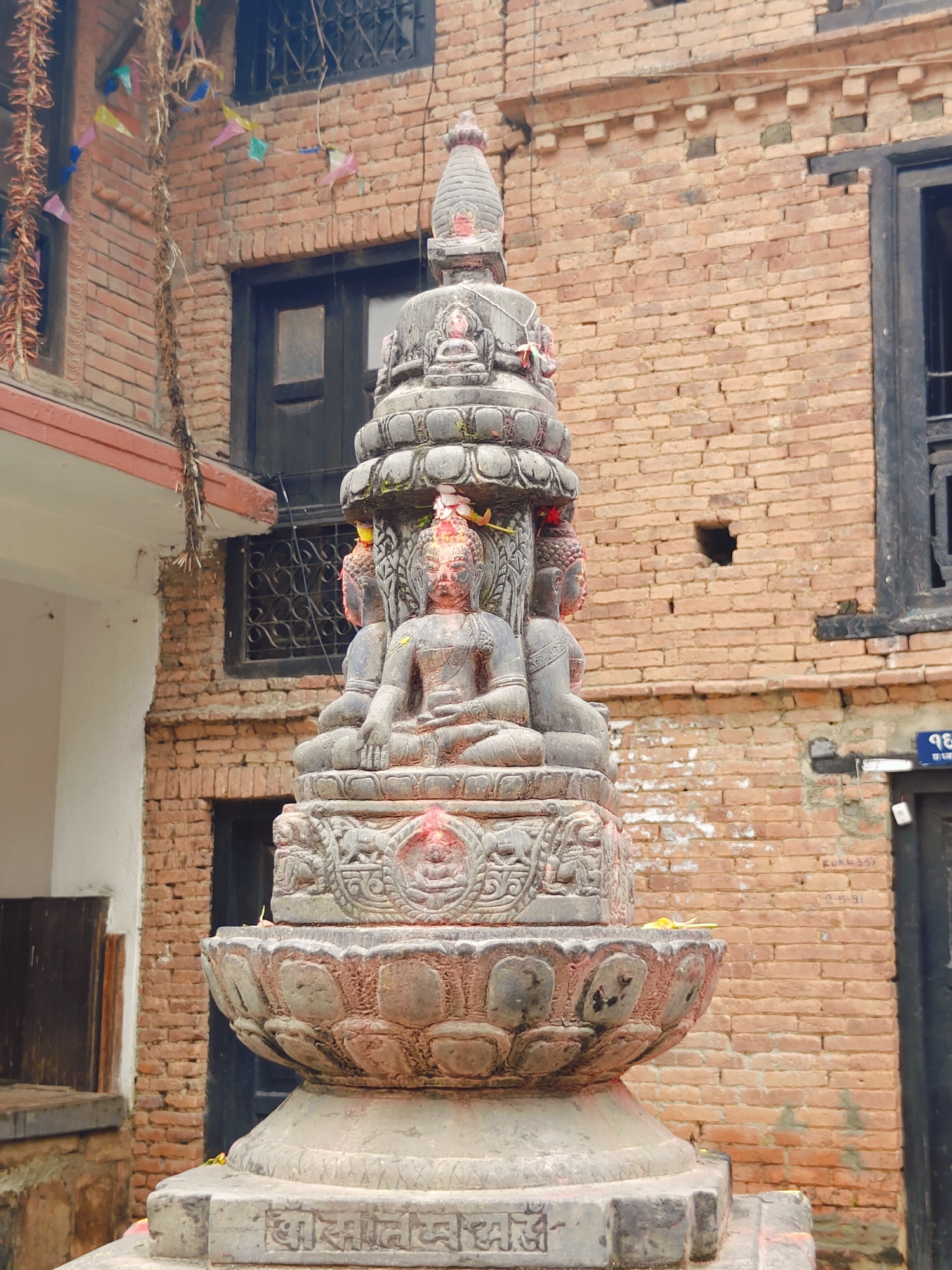 The image shows a stone stupa with multiple Buddha statues in Kirtipur, Nepal. The stupa is intricately carved with floral motifs and sits on a lotus-shaped base. The statues of Buddha are seated in meditative poses. In the background, a brick building with dark windows and a few prayer flags are visible. The overall setting appears to be an outdoor religious site in a city.