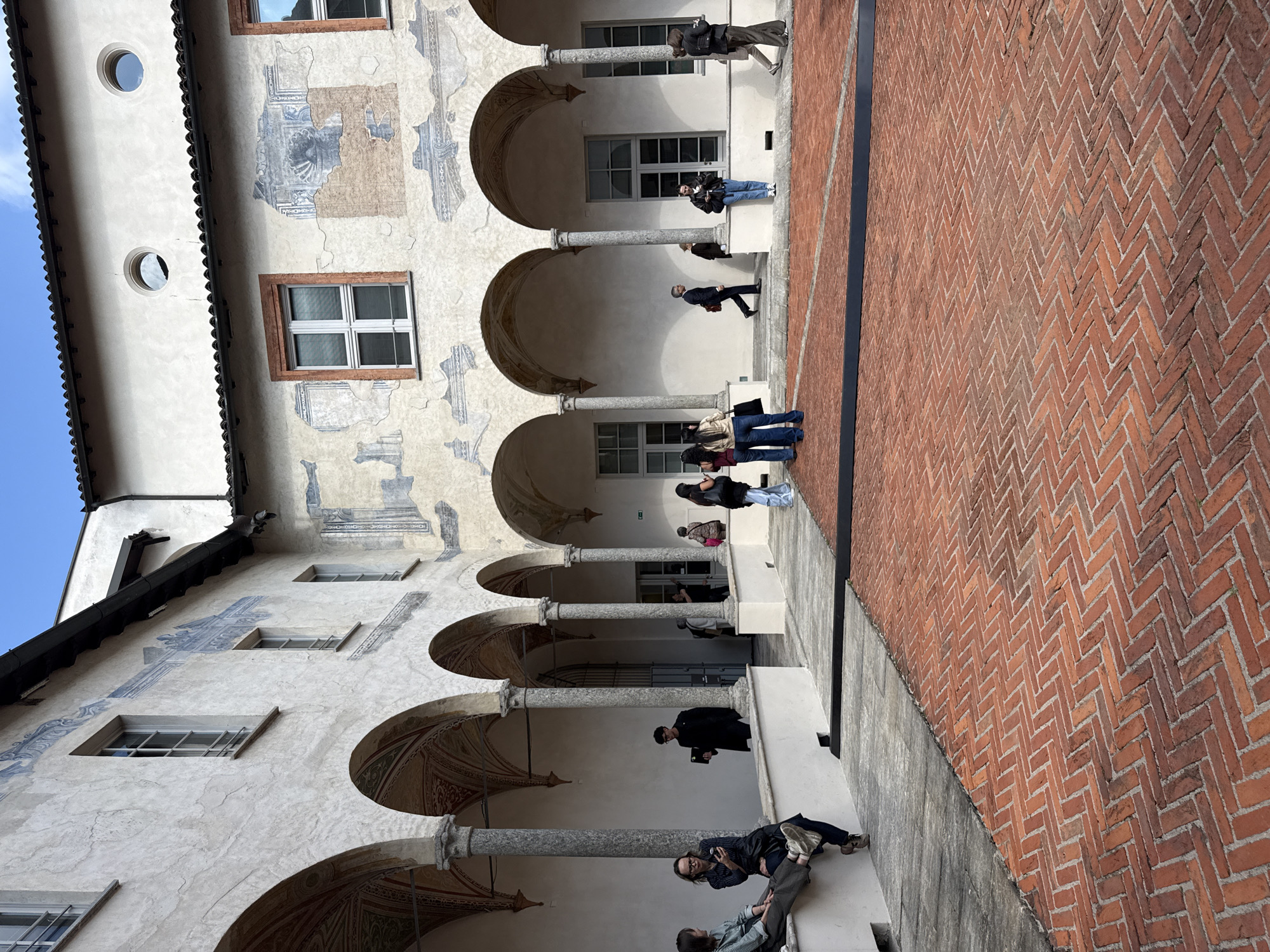 This media file captures a bustling courtyard or cloister of an old building, likely a historical institution or palace, in Milan, Italy, under a clear blue sky.
The scene features a long arcade or portico extending along the right side of the image, defined by a series of arched openings supported by stone pillars. The walls of the building are a light-colored stucco, adorned with extensive areas of faded frescoes and murals, depicting intricate patterns and architectural elements in muted tones of blue, gray, and brown. Above the arcade, several rectangular windows with white frames are visible, along with two distinct circular windows (oculi) on the upper left section of the facade. The inner surfaces of the arches also show remnants of decorative painting.
The ground of the courtyard is paved with reddish-brown bricks laid in a distinctive herringbone pattern, with a dark, possibly concrete or stone, strip running parallel to the building.
Numerous people, mostly adults, are scattered throughout the courtyard, engaged in various leisurely activities. In the foreground, a group is seated or lounging on a low wall or bench, with one person looking at a smartphone. Under the arches of the arcade, several individuals are standing or walking; one person appears to be holding a tablet or notebook, while others observe their surroundings or interact. Further along the arcade, more people are seen strolling or standing.
The overall ambiance suggests a public space, possibly a museum, gallery, or educational institution, where visitors are exploring or relaxing. No prominent or legible text is visible in the image.