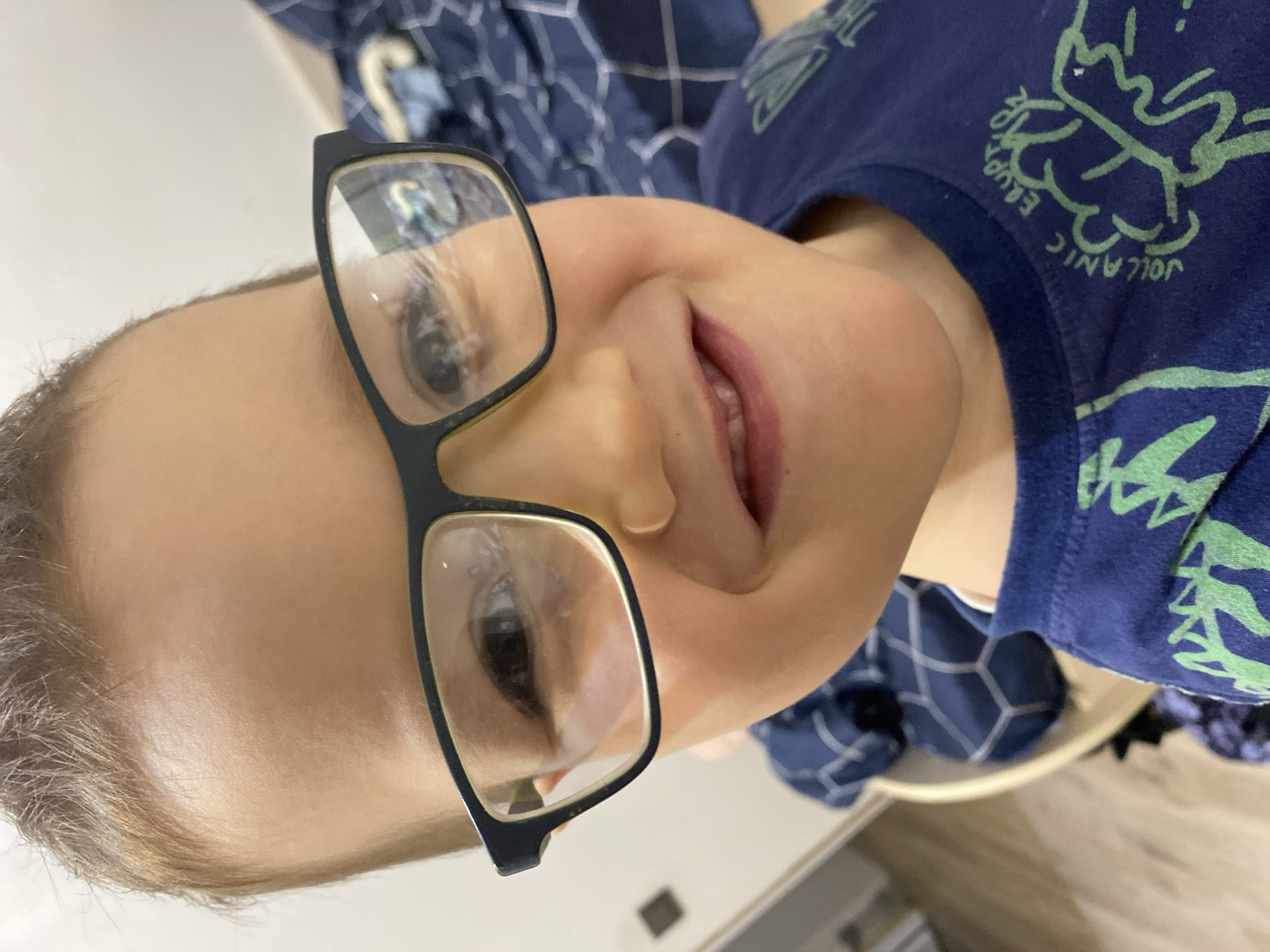 A young child with short, light brown hair is wearing glasses and a blue shirt with green dinosaur-like designs. The child is smiling slightly, and their face is angled towards the camera. The background includes a blue and white patterned fabric and a light-colored surface that appears to be a table or countertop. The location is identified as Chaikovsky, Russia.