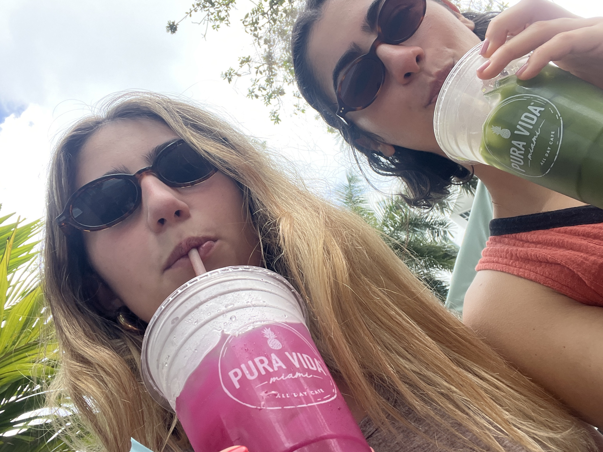 Two women, likely in their 20s, are shown from a low-angle perspective, holding drinks from Pura Vida Miami, a restaurant chain with multiple locations in Miami Beach, Florida, United States of America. The woman on the left has long blonde hair and is wearing sunglasses while drinking from a pink-colored beverage using a straw. The woman on the right has dark hair, is also wearing sunglasses, and is drinking from a green-colored beverage. Green foliage is visible in the background, suggesting an outdoor setting. The sky is partly cloudy.