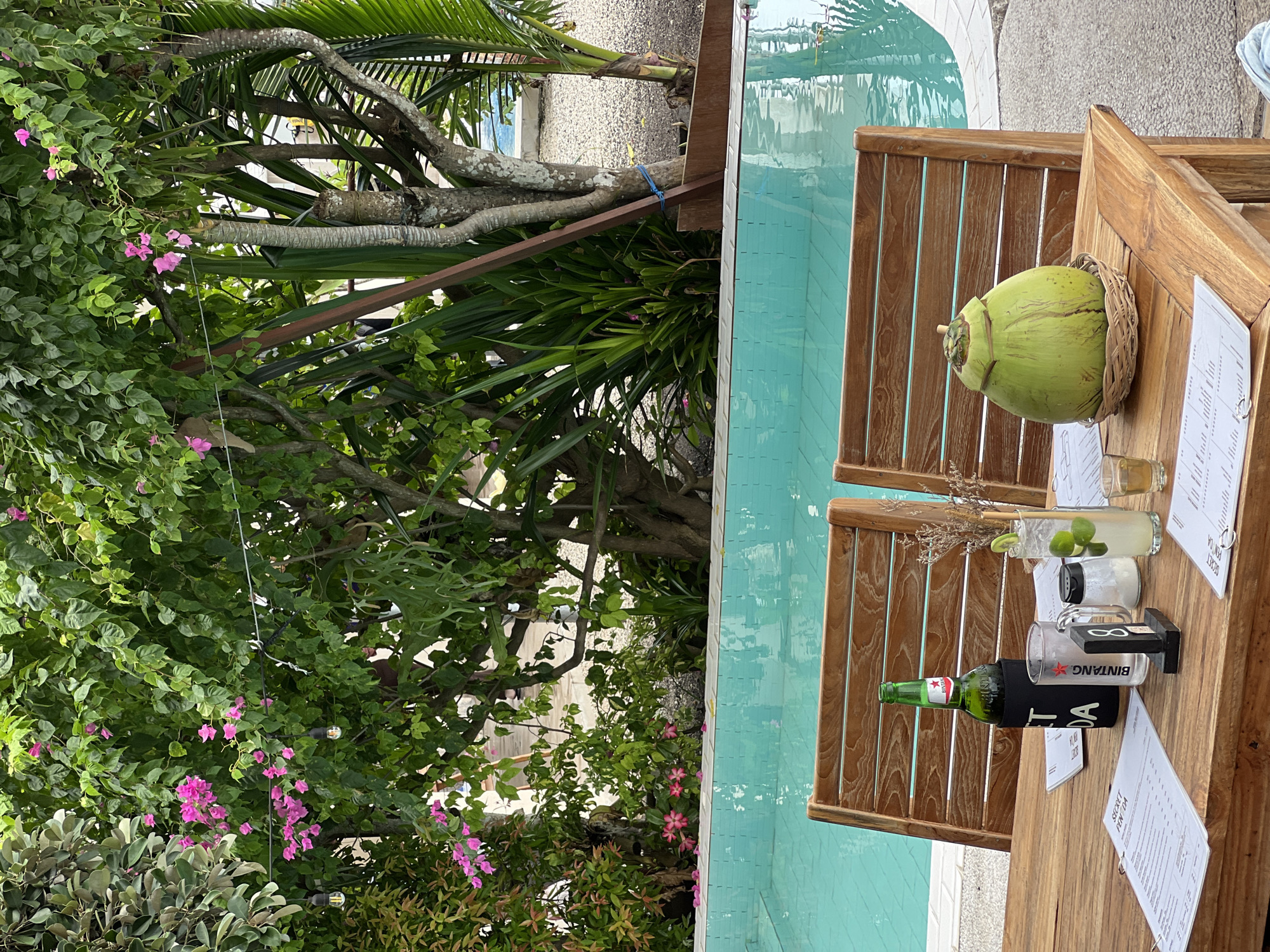 The image displays a vibrant outdoor scene, likely a resort or cafe, in a tropical setting. The main subject is a rustic wooden table positioned alongside a light blue swimming pool. On the table, several refreshing beverages are arranged, suggesting a leisurely moment.
Visible items on the table include a large green young coconut served in a woven basket, a green glass bottle of Bintang beer in a black coozie, and a tall clear glass containing a drink with ice and lime slices, possibly a cocktail or soda water with lime. Another smaller glass with a dark liquid is also present, along with a salt shaker and a small black rectangular object, possibly a stand or coaster. Several white papers, likely menus or order forms, are scattered on the table, one clearly showing "SECRET PENIDA" as part of its text. Two wooden chairs or stools are partially visible next to the table.
The environment is lush and verdant, dominated by dense tropical foliage. Bright pink bougainvillea flowers cascade from overhead, adding a splash of color against the green leaves. Large palm fronds and thick tree trunks are prominent in the background, further emphasizing the tropical ambiance. The swimming pool features light blue tiles and clear water, reflecting the bright outdoor light.
The scene conveys a sense of relaxation and vacation, likely during the daytime, given the bright lighting and the nature of the outdoor setting by the pool. The presence of Bintang beer and the text "SECRET PENIDA" strongly suggest the location is in Nusa Penida, Indonesia. No people are visible in the frame.
