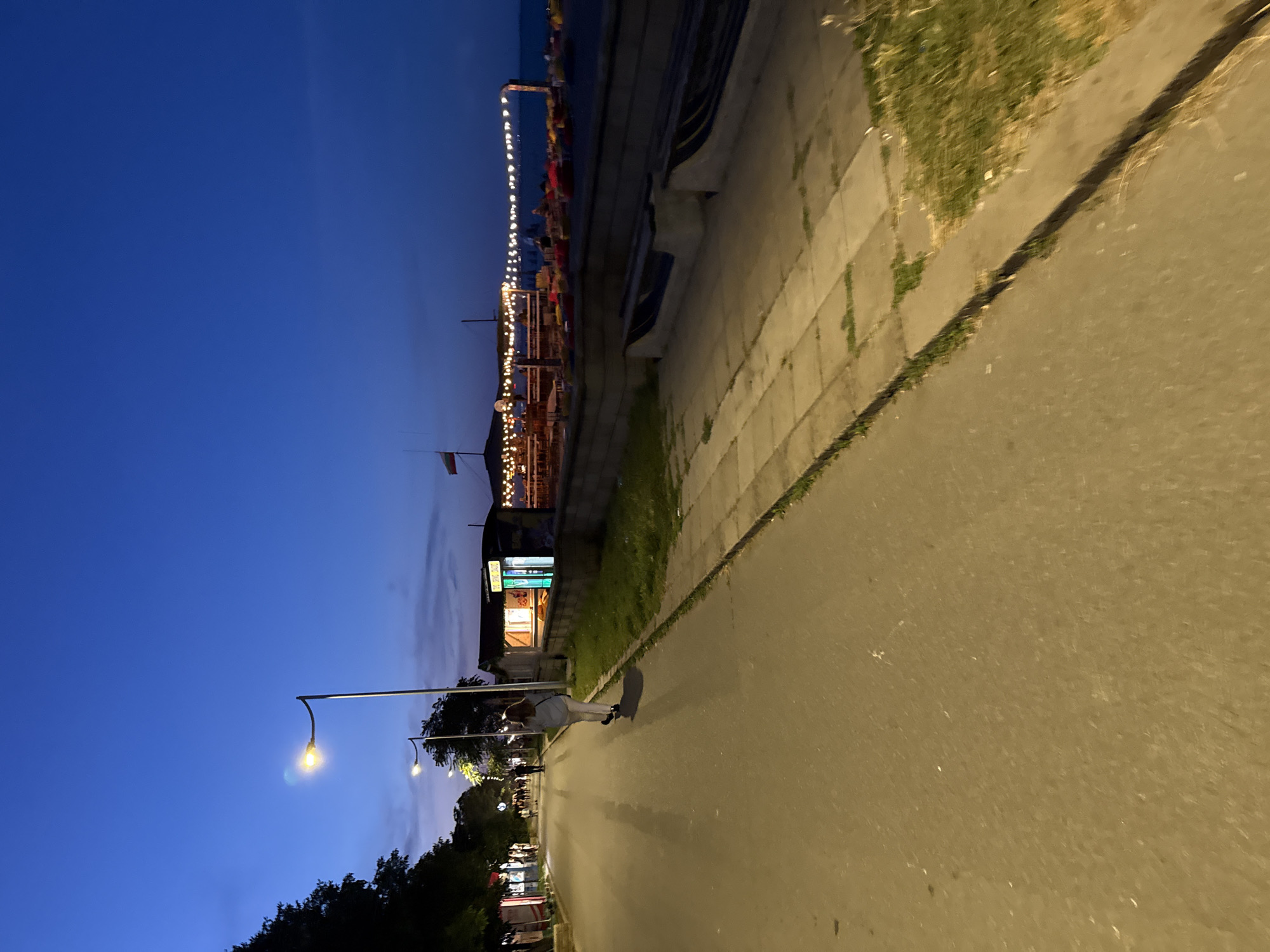 The image depicts a street scene at dusk in Burgas, Bulgaria. A person is walking away from the camera on a paved road. To the right of the road, there's a sidewalk with concrete slabs and grass. Beyond the sidewalk, a row of restaurants and bars with illuminated signs and string lights are visible along a waterfront area. The sky is a deep blue, transitioning from twilight. In the distance, the sea or a large body of water can be seen. Streetlights illuminate the pathway. The overall atmosphere is calm and relaxed, suggesting a popular evening gathering spot.