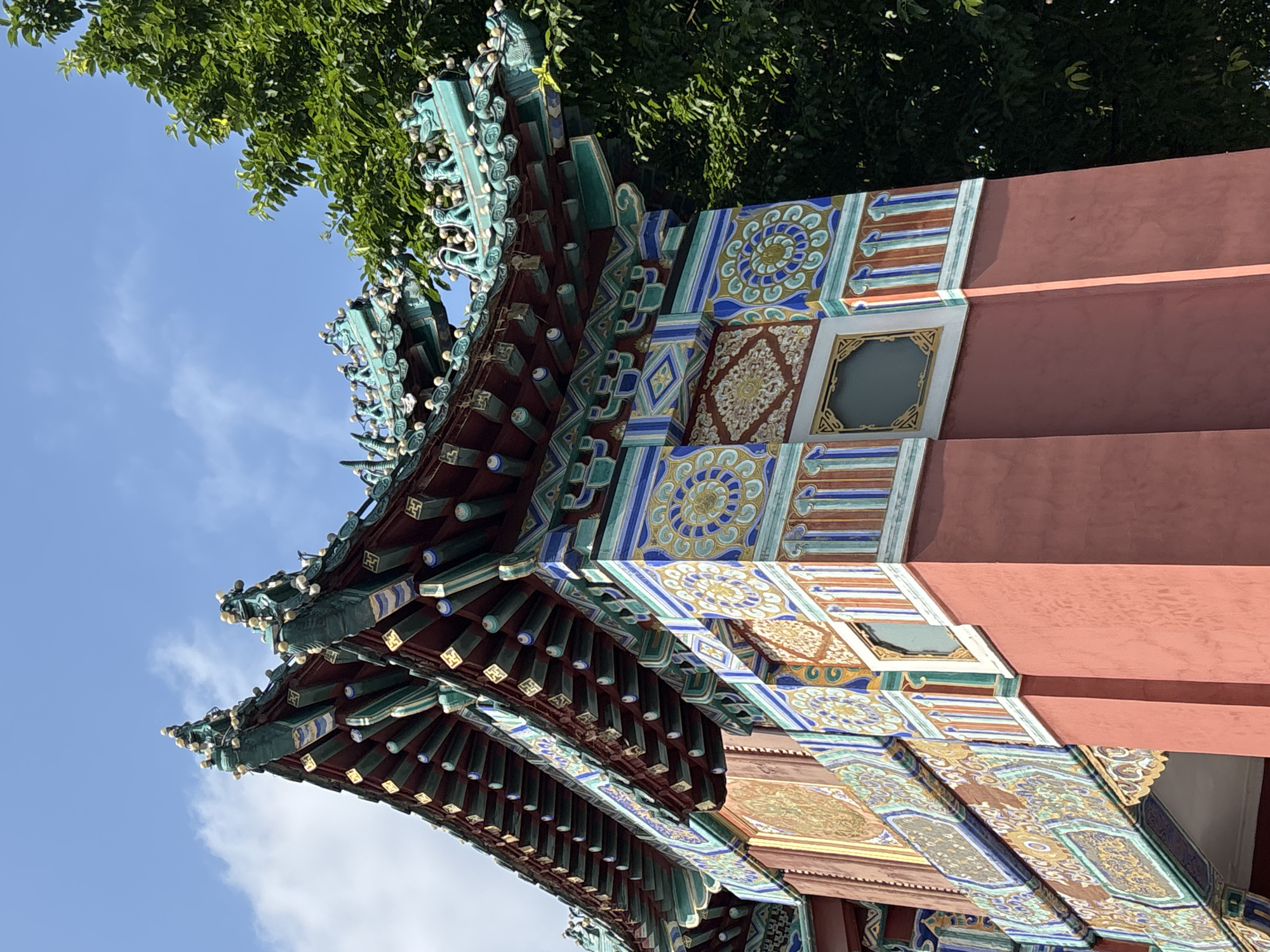 This is a close-up, low-angle shot of the ornate roof and upper portion of a vibrant, traditional Chinese building. The building features a multi-tiered, dark red roof with green, turquoise, and gold decorative trim, including figures of what appear to be mythical creatures or statues along the eaves. The supporting structure is a deep red, with intricate patterns of yellow, blue, and white adorning its surface in geometric and floral motifs. Visible below the roofline, there are sections of gold ornamentation. The background consists of a bright blue sky with scattered white clouds and the lush green foliage of a large tree on the right side of the frame. The image appears to be taken during the daytime. The style of architecture suggests it could be a temple, palace, or historical gate. Given the mention of Yuzhong District, China, this is likely a cultural or historical landmark within that region. There are no people or visible actions in the image.