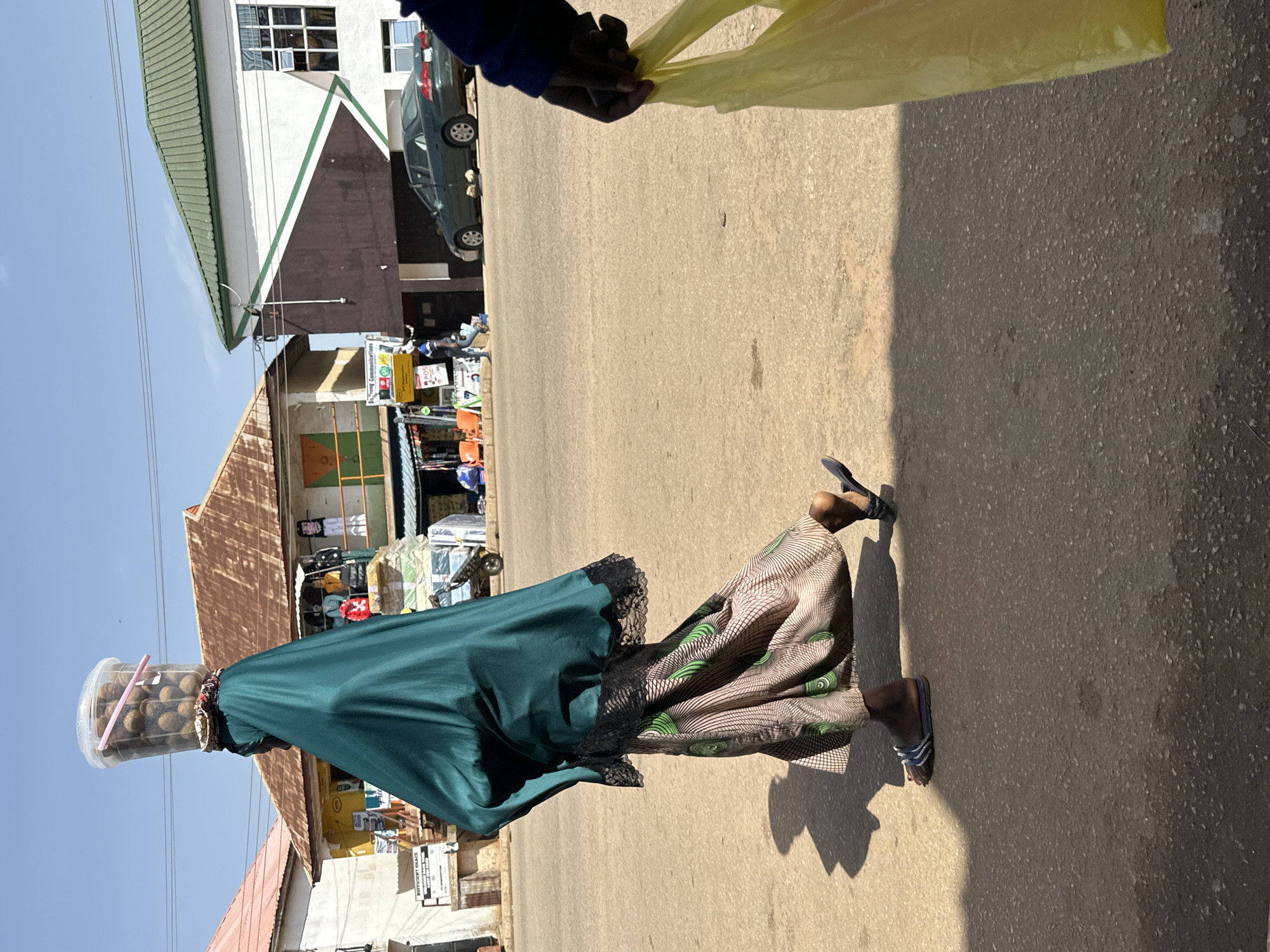 In Bukuru, Nigeria, a person walks on a paved street, their head balanced with a large plastic container filled with spherical items, possibly nuts or seeds, secured by a pink stick. They are draped in a dark green fabric, possibly a hijab or veil, with a lace trim at the edge, and wear a patterned skirt. The person is also wearing sandals. A yellow plastic bag is visible in the foreground, held by a hand. Buildings line the street, some with corrugated iron roofs, others with modern facades and green trim. A dark green car is parked on the street. The environment appears to be a bustling marketplace or street with various stalls and people visible in the background. The lighting suggests a bright, sunny day, casting distinct shadows on the ground.