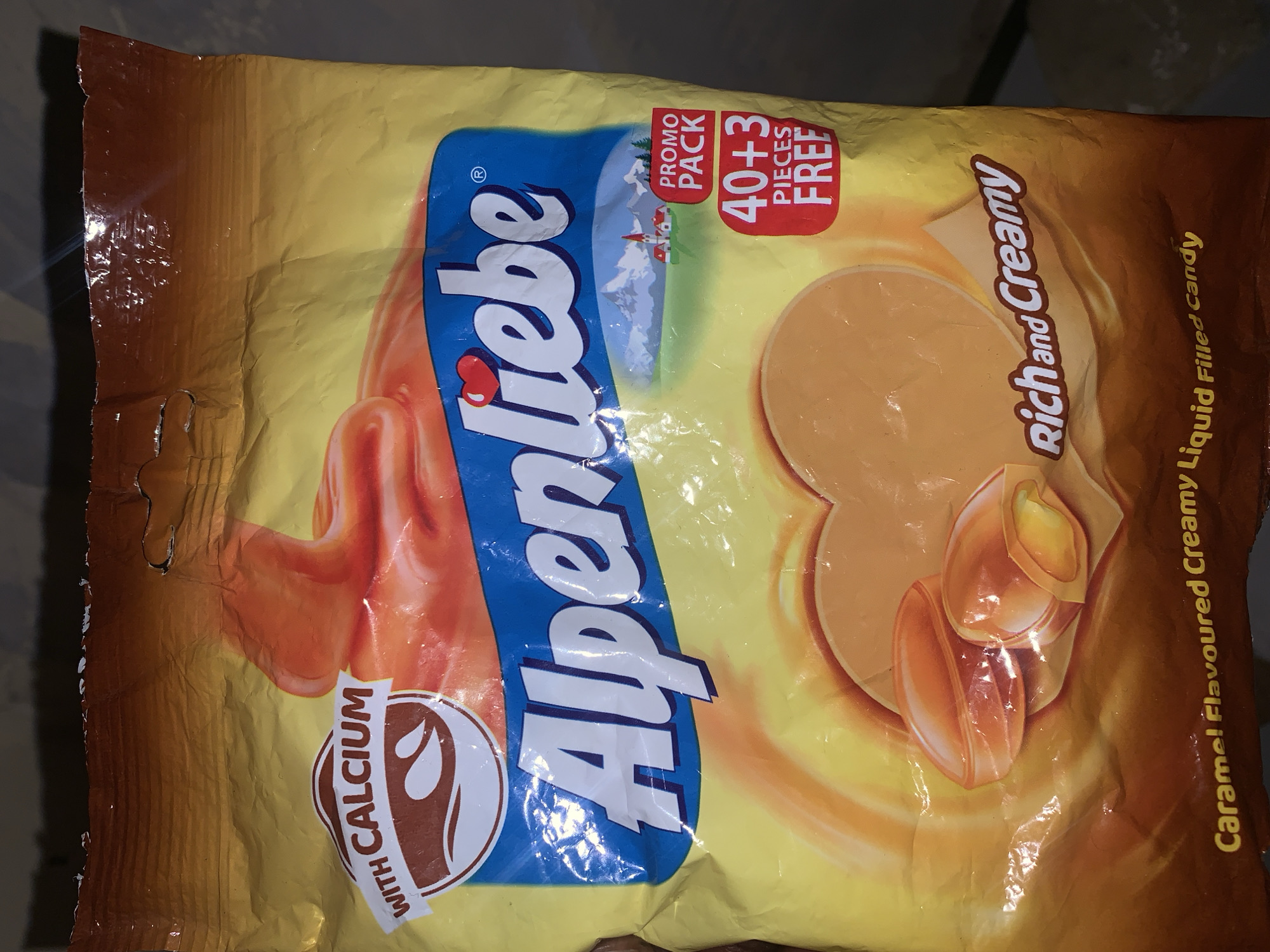 The image shows a promotional packet of Alpenliebe brand caramel-flavored candy. The flexible plastic packaging is primarily yellow and brown, oriented diagonally across the frame against a dark, indistinct background, suggesting an indoor setting.
The prominent brand name "Alpenliebe" is displayed in white text with a red heart over the 'i', set against a curved blue banner that illustrates a snow-capped mountain range and a small village. A red rectangular label on the upper right side announces a "PROMO PACK" offering "40+3 PIECES FREE."
Further details on the packet include the phrases "Rich and Creamy" in a script font, and "WITH CALCIUM" inside a circular emblem featuring a milk drop. The full product description, "Caramel Flavoured Creamy Liquid Filled Candy," is printed vertically along the right side. The design also features realistic depictions of the caramel candies, some showing their creamy liquid filling. The packet has some visible creases and reflections, typical of plastic packaging. No people or specific location cues are visible.