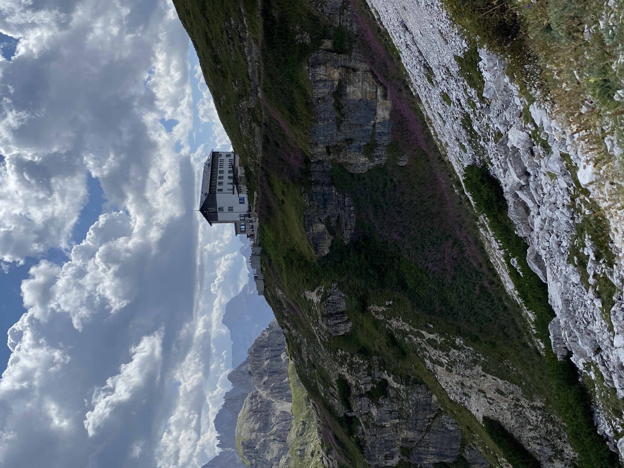 A white and dark building with multiple windows stands on a green, rocky mountainside under a cloudy sky. The building appears to be a lodge or hotel, with a dark roof and a series of white sections. It is situated on a ledge overlooking a steep slope covered in green vegetation and scattered purple flowers. In the background, rugged mountain peaks are visible, shrouded in a light haze. The foreground is dominated by a rocky, scree-covered slope, with patches of green shrubs and smaller rocks. There are no visible people or any actions taking place in the image. The scene is a panoramic view of a mountain landscape, suggesting a remote, natural setting. The location is identified as Auronzo di Cadore, Italy.