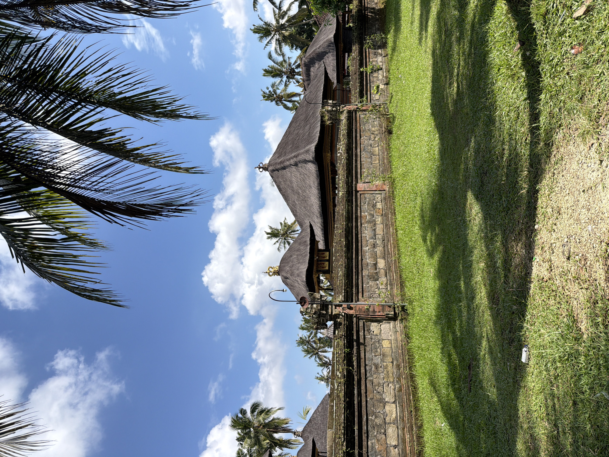 The image depicts a clear blue sky with scattered white clouds, partially obscured by the fronds of palm trees in the foreground. Below the sky, a stone wall, weathered and overgrown with moss and vegetation, separates a well-maintained grassy lawn from a series of traditional Balinese buildings. These buildings feature thatched roofs with ornate crests, indicative of a temple or royal complex. Palm trees are visible in the background, dotting the landscape. The ground in the foreground is covered in lush green grass, with patches of gravelly soil. The lighting suggests a sunny day, casting shadows from the palm trees onto the lawn. The location is identified as Tampaksiring, Indonesia, a region known for its historical and cultural sites. No people are visible in the scene.