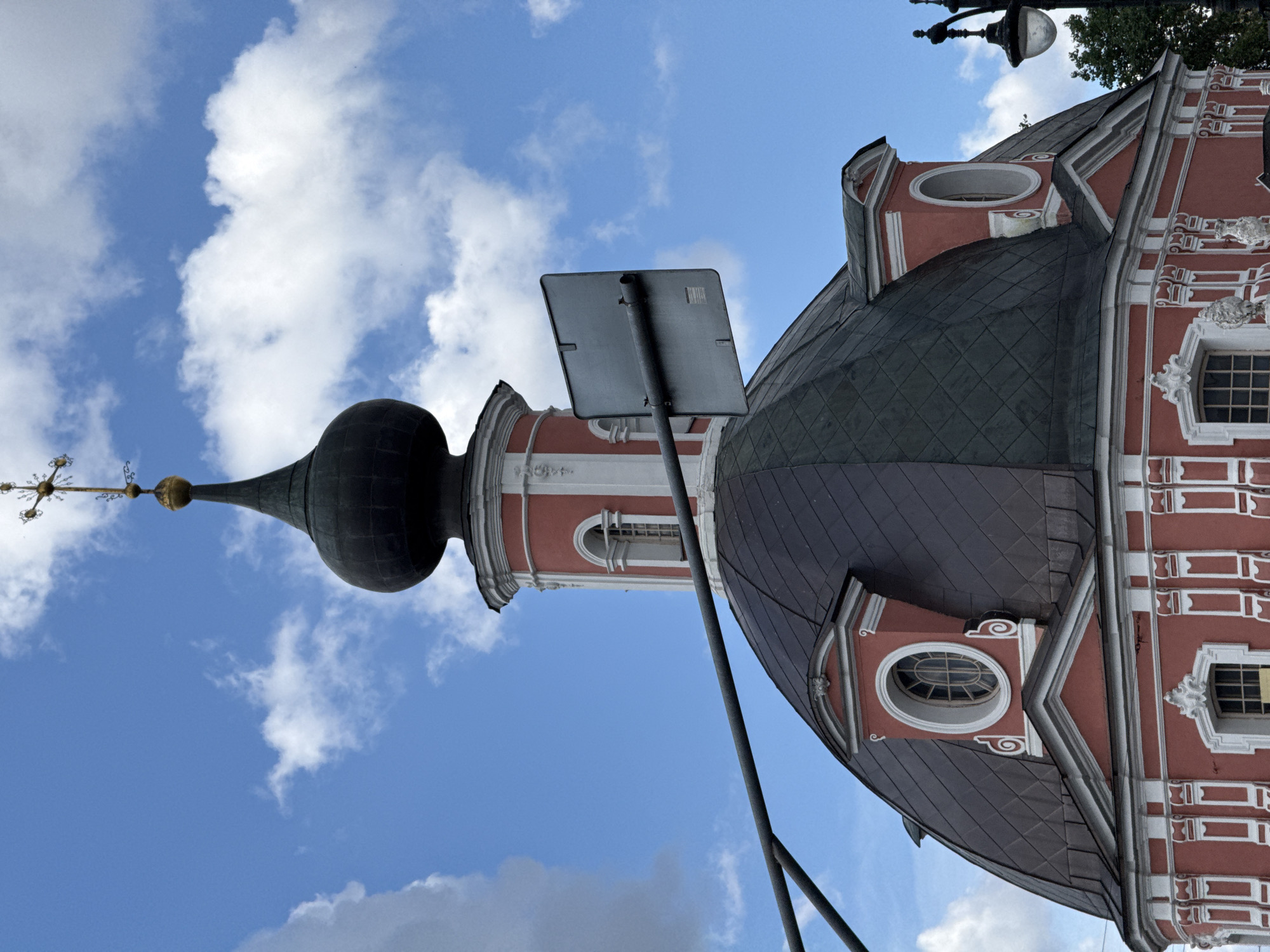 The image shows the dome and upper sections of the Church of the Resurrection of Christ in Kadashi, Moscow, Russia. The building is painted a distinct coral pink with white decorative elements. The main dome is dark, possibly slate or copper, with a rounded shape, and features oval windows with white trim. Above this dome is a smaller, red and white tower, also topped with a dark, bulbous dome, from which a golden cross extends into the bright blue sky dotted with white clouds. A horizontal metal pole and a square, grey sign are visible in the foreground, partially obscuring the building. To the right, a traditional black lamppost with a lantern is visible, as are the tops of trees and a sliver of the sky. The overall scene depicts an architectural detail of a historic religious building under a partly cloudy sky.