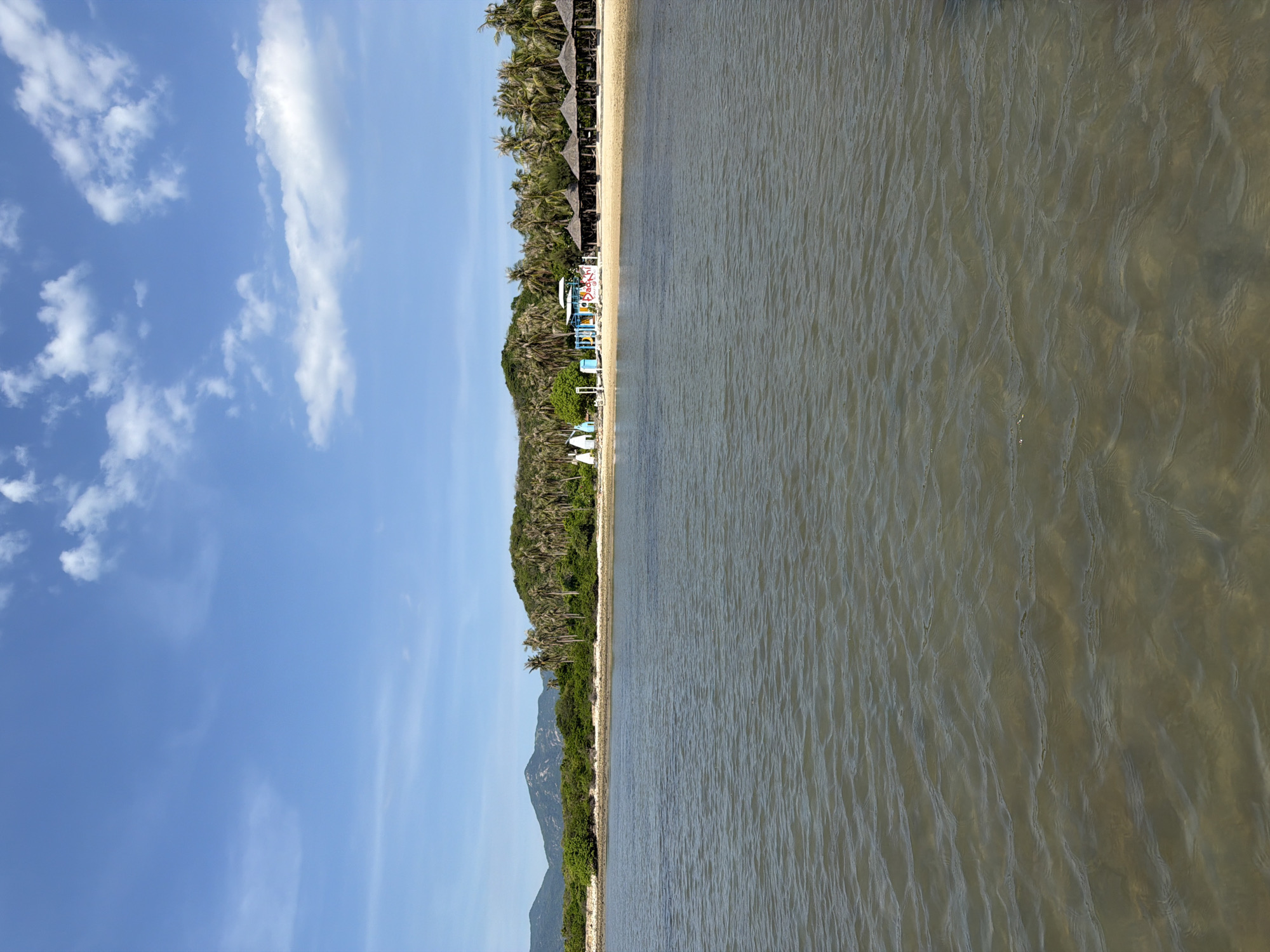 A serene coastal scene unfolds with the water in the foreground, a tranquil expanse of muted brown with gentle ripples reflecting the sunlight. The background reveals a sandy beach lined with a row of thatched-roof umbrellas, suggesting a resort or a recreational area. Beyond the beach, a dense cluster of palm trees forms a lush backdrop, merging with rolling green hills and distant mountains under a bright blue sky dotted with wispy white clouds. A vibrant, multi-colored structure, possibly part of a playground or amenity, stands on the beach near the water's edge. The overall impression is one of a peaceful, tropical holiday destination. The location is the coast of Ninh Ích, Vietnam.