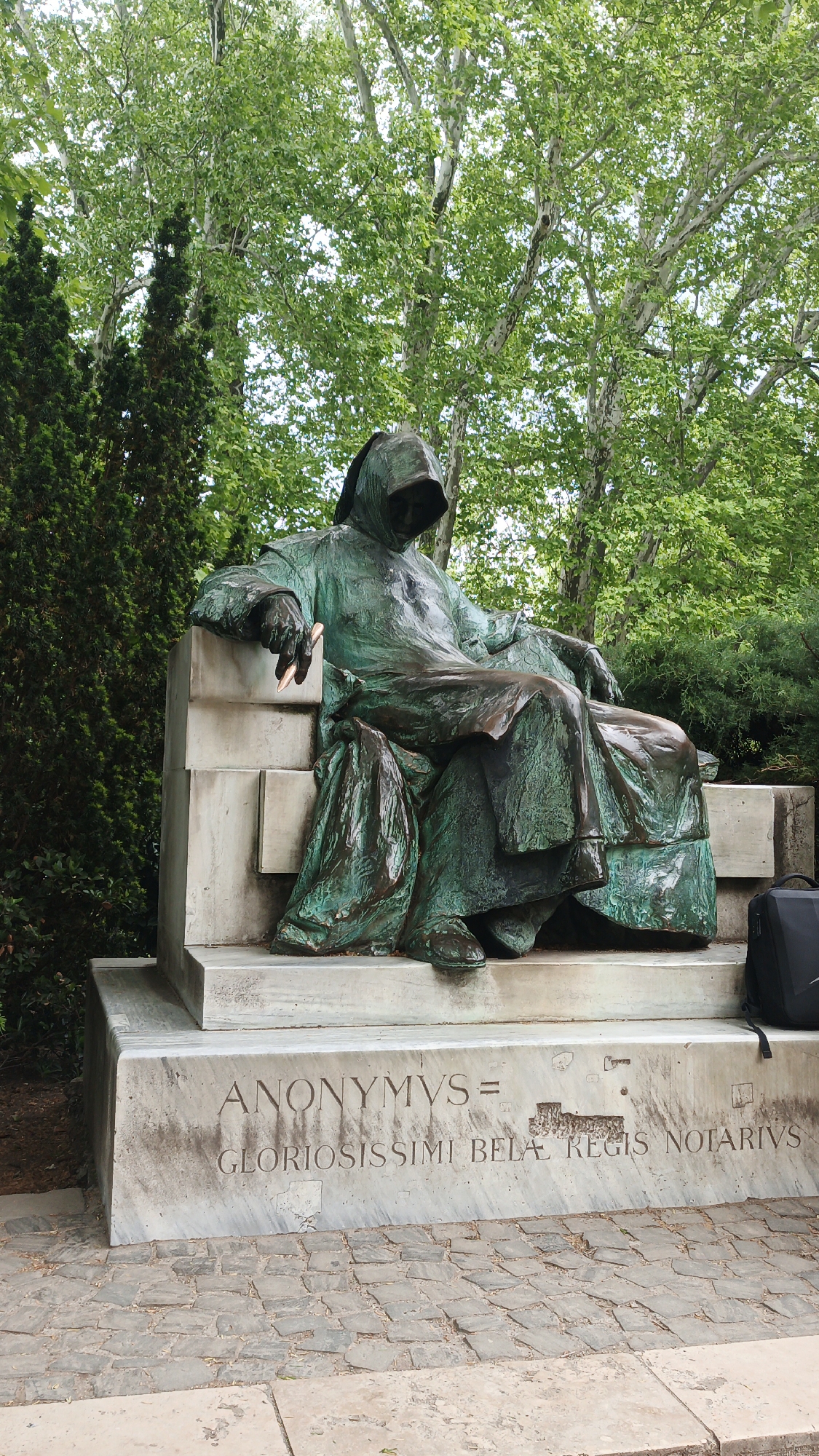 The image shows the statue of Anonymus, an anonymous chronicler and notary of King Béla III of Hungary in the 12th century. The bronze statue depicts a hooded figure seated on a stone pedestal. He is draped in robes and holds a quill in his left hand. The pedestal has the inscription "ANONYMVS = GLORIOSISSIMI BELÆ REGIS NOTARIVS". A black backpack sits behind the pedestal. The statue is located in Városliget (City Park), Budapest, Hungary. The surrounding environment consists of trees and shrubs. The ground in front of the statue is paved with stones.