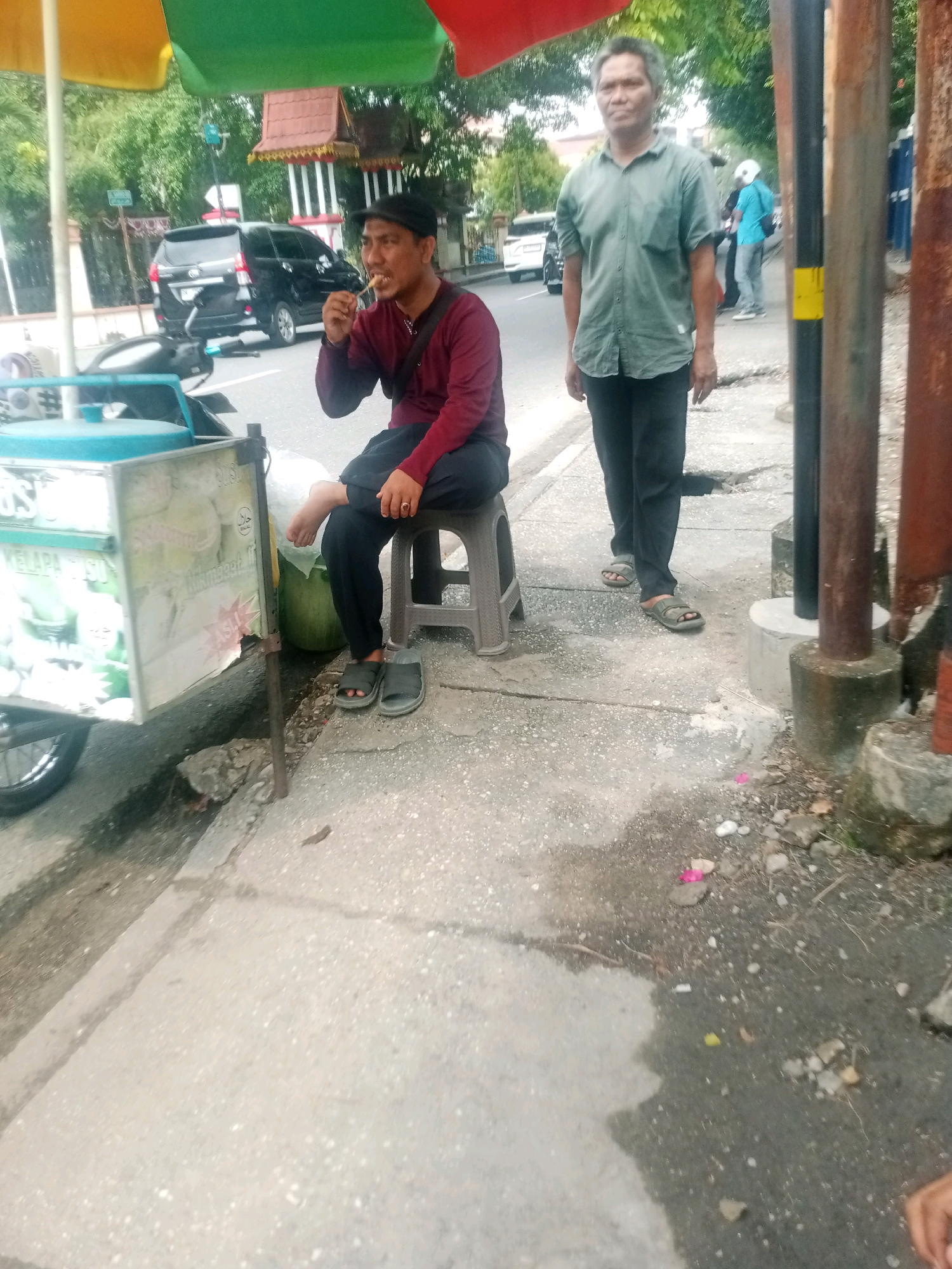The image depicts a street scene in Pekanbaru, Indonesia, under a bright, overcast sky. In the foreground, a street vendor sits on a gray plastic stool, eating from a skewer. He is wearing a maroon long-sleeved shirt, black pants, a black cap, and dark gray sandals. Next to him is a cart, likely for selling food or drinks, with a sign on its side. To the right of the vendor, another man stands, wearing a green short-sleeved collared shirt and black pants, also in dark gray sandals. He appears to be looking towards the viewer. Behind them, a black SUV is parked on the road, with other vehicles and trees visible in the background. The sidewalk is paved with concrete, and there are several cylindrical concrete structures and metal posts along the side of the road. Debris, including small stones and a few pink flower petals, is scattered on the ground. The overall impression is that of a typical urban street environment.