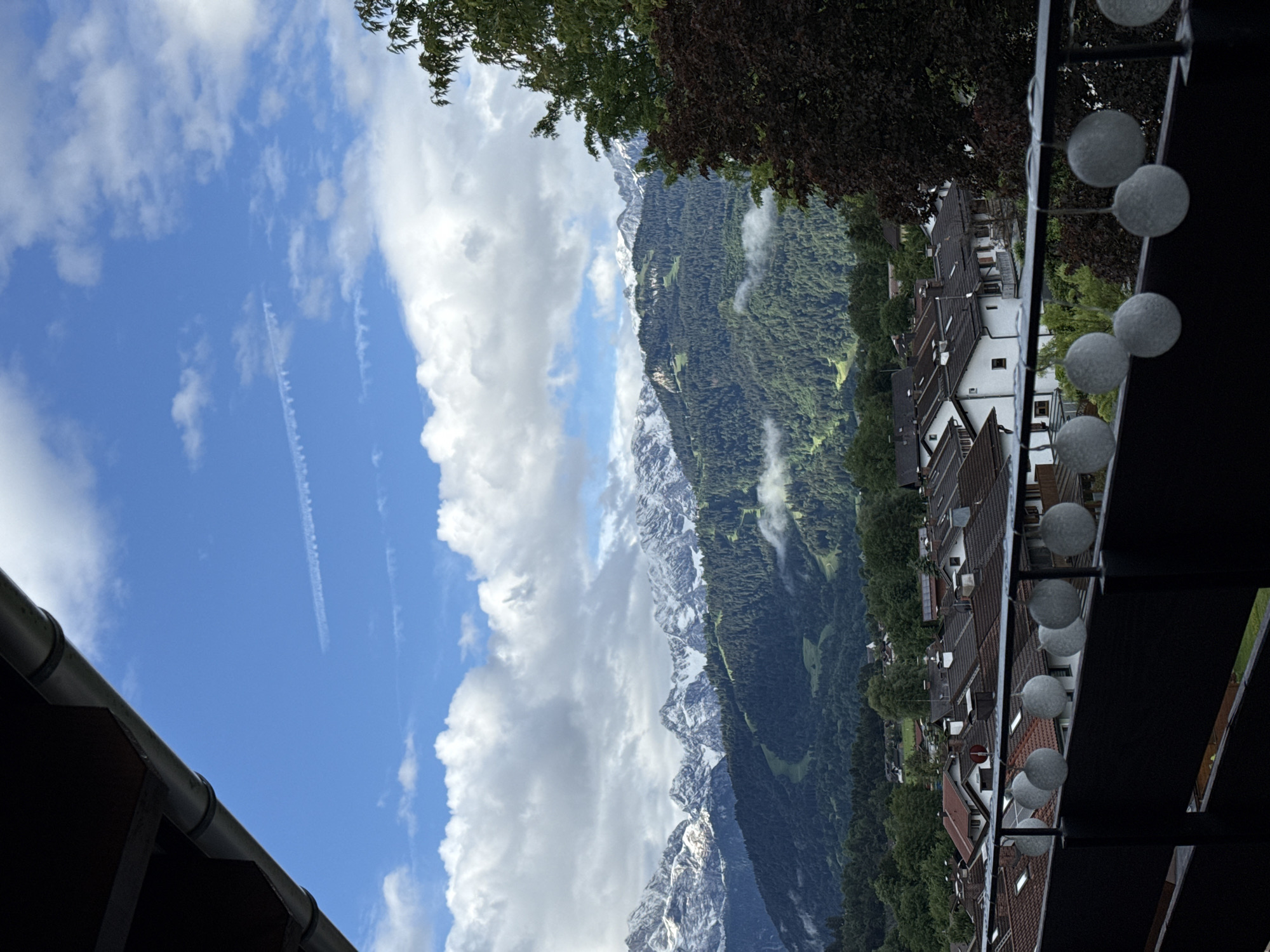 The image depicts a view from a balcony overlooking a village nestled in the foothills of snow-capped mountains. The foreground features a dark railing with a string of circular, globe-shaped lights. Beyond the railing, a dense arrangement of residential buildings with terracotta roofs and white facades is visible, interspersed with trees. Further in the distance, a vast expanse of verdant, forested mountains rises, culminating in rugged, snow-dusted peaks under a bright blue sky with scattered white clouds. Faint contrails streak across the sky, suggesting the presence of aircraft. The overall scene suggests a picturesque location in the Bavarian Alps, likely in or near Garmisch-Partenkirchen, Germany, given the characteristic alpine architecture and mountain landscape. No people or specific actions are depicted in the image.