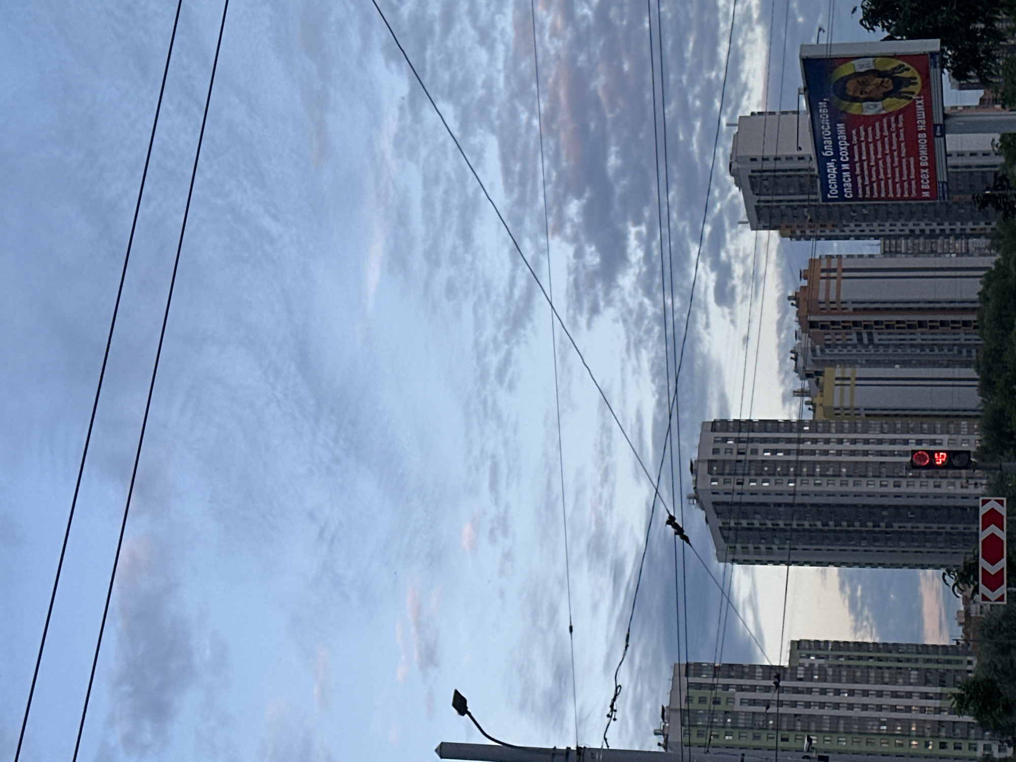 The image shows a view of residential buildings and a billboard against a cloudy sky in Saint Petersburg, Russia. Multiple power lines crisscross the sky. On the right side of the image, a billboard features a religious text in Cyrillic that reads "Господи, благослови, спаси и сохрани и всех воинов наших!" which translates to "Lord, bless, save and protect, and all our warriors!". The billboard also has an image of a man's face. In the foreground, traffic signals and road signs are visible. The buildings are modern high-rise apartments. The overall atmosphere is that of a city street during twilight.