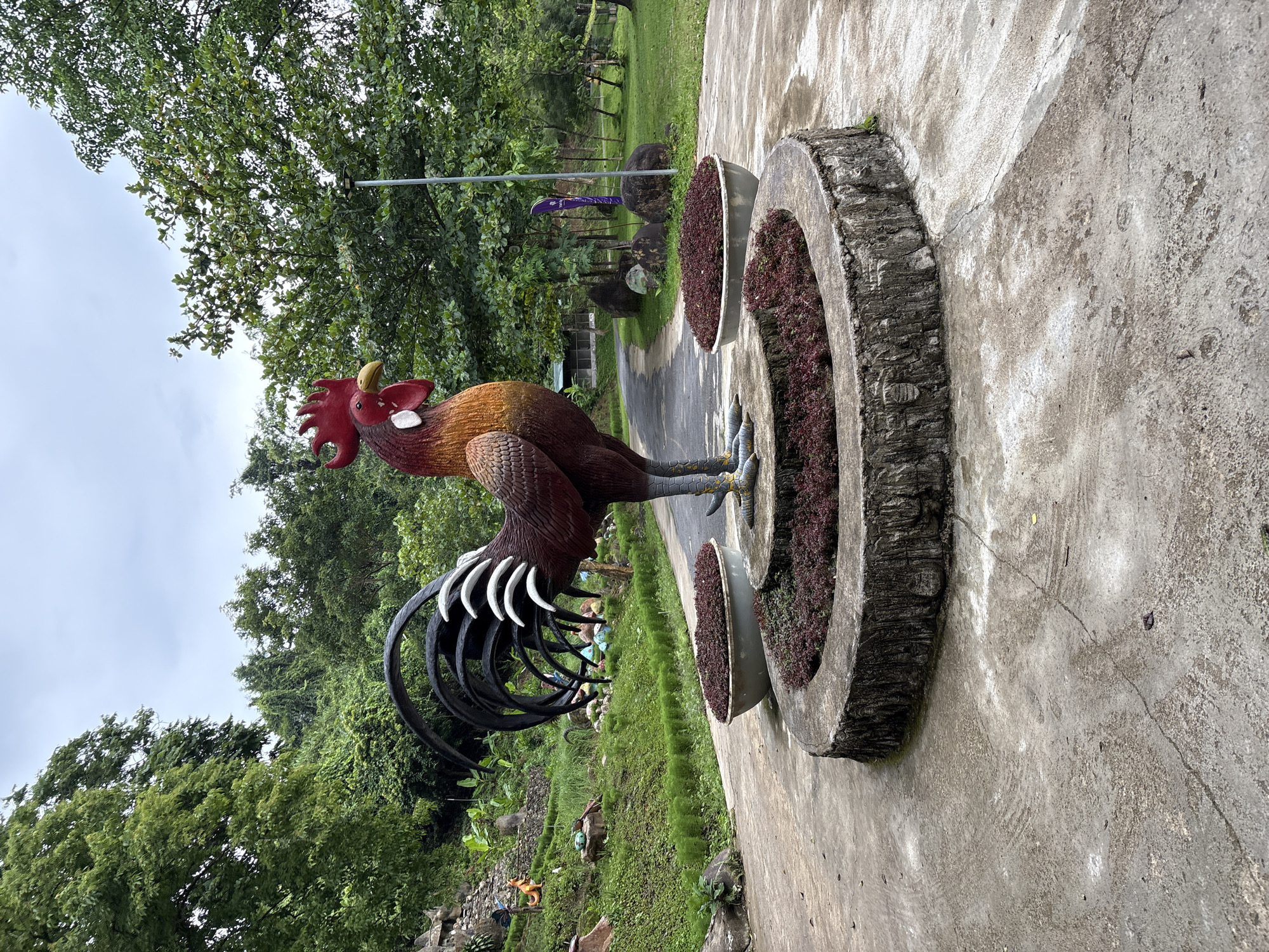 This is a frontal view of a large rooster statue in a park-like setting, likely in Khanh Vinh, Vietnam. The statue is made of painted fiberglass or similar material, featuring a reddish-brown body with yellow accents on its wings and neck. It has a bright red comb and wattle, a yellow beak, and grey legs with prominent claws. The rooster stands on a circular, raised platform with a textured, wood-like finish, which is surrounded by a low, circular garden bed filled with dark red plants.
To the left of the statue, a sloped, grassy area is visible, adorned with smaller decorative statues of animals, including what appears to be a fox-like figure, a dinosaur, and other indistinct figures. Further up the slope, there are rocks and lush green vegetation.
The statue is positioned on a paved pathway that curves into the background. This pathway leads through a green area with trees and grass. In the mid-ground, a light pole with a black fixture stands near the pathway, and in the background, more trees and a cloudy sky are visible. The overall environment suggests an outdoor recreational space or a garden. The paving is a light gray concrete, showing some wear and tear, with visible cracks and stains. The lighting is diffused, consistent with an overcast day. There are no people visible in the image.