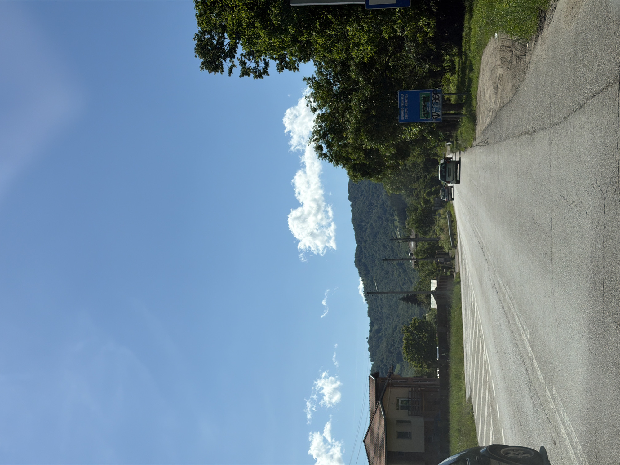 The image depicts a rural road scene in Beli Osam, Bulgaria, under a clear blue sky with scattered white clouds. A paved road, with visible white lane markings and some wear and tear, stretches into the distance. On the left side of the road, there is a building with a tiled roof and a balcony. Further back, a treelined hillside forms the backdrop. On the right side of the road, a dense cluster of green trees is prominent. Attached to a pole within the trees is a blue sign with white lettering that reads "ШИПКООВО-РИБАРИТСА SHIPKOVO-RIBARTSI" indicating a place name. Below this sign, there is another sign with green and white text and a graphic. A few cars are visible on the road, with one car in the foreground on the left, partially cropped, and two cars further down the road, moving away from the viewer. The environment appears to be a warm, sunny day.