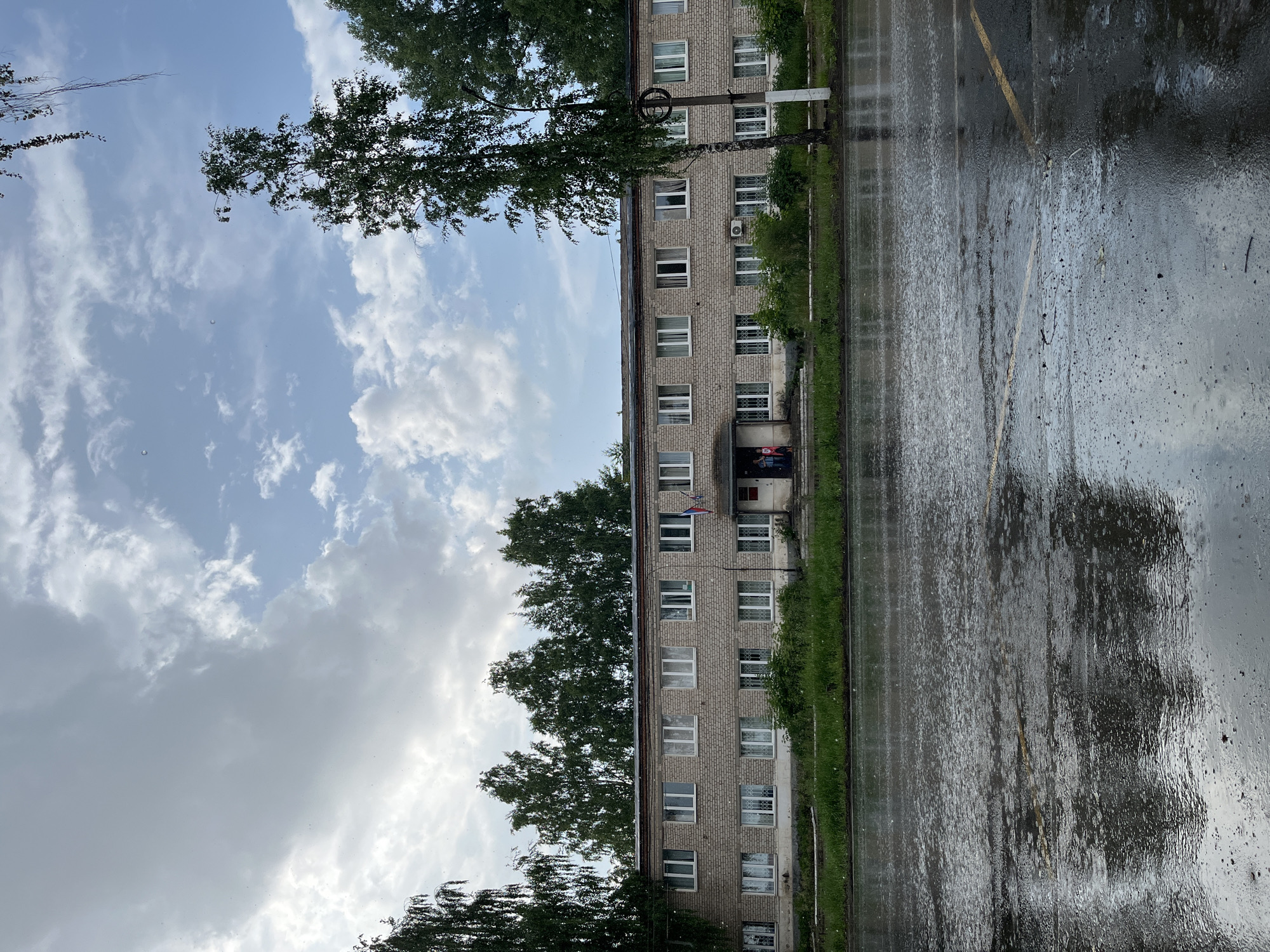 The image captures a rainy day scene in the city of Chaikovsky, Russia, likely in front of a residential or administrative building. The building is a two-story brick structure with multiple windows on each floor, some of which have white curtains. A small group of people, possibly two children and an adult, are visible near the entrance, which has a small awning. A Russian flag is mounted on the facade of the building. Trees line the front of the building, and a grassy area separates it from the street. The street in the foreground is wet and reflective, with visible rain streaks, indicating active rainfall. The sky is overcast with a mix of clouds. A lamppost stands to the right of the building.