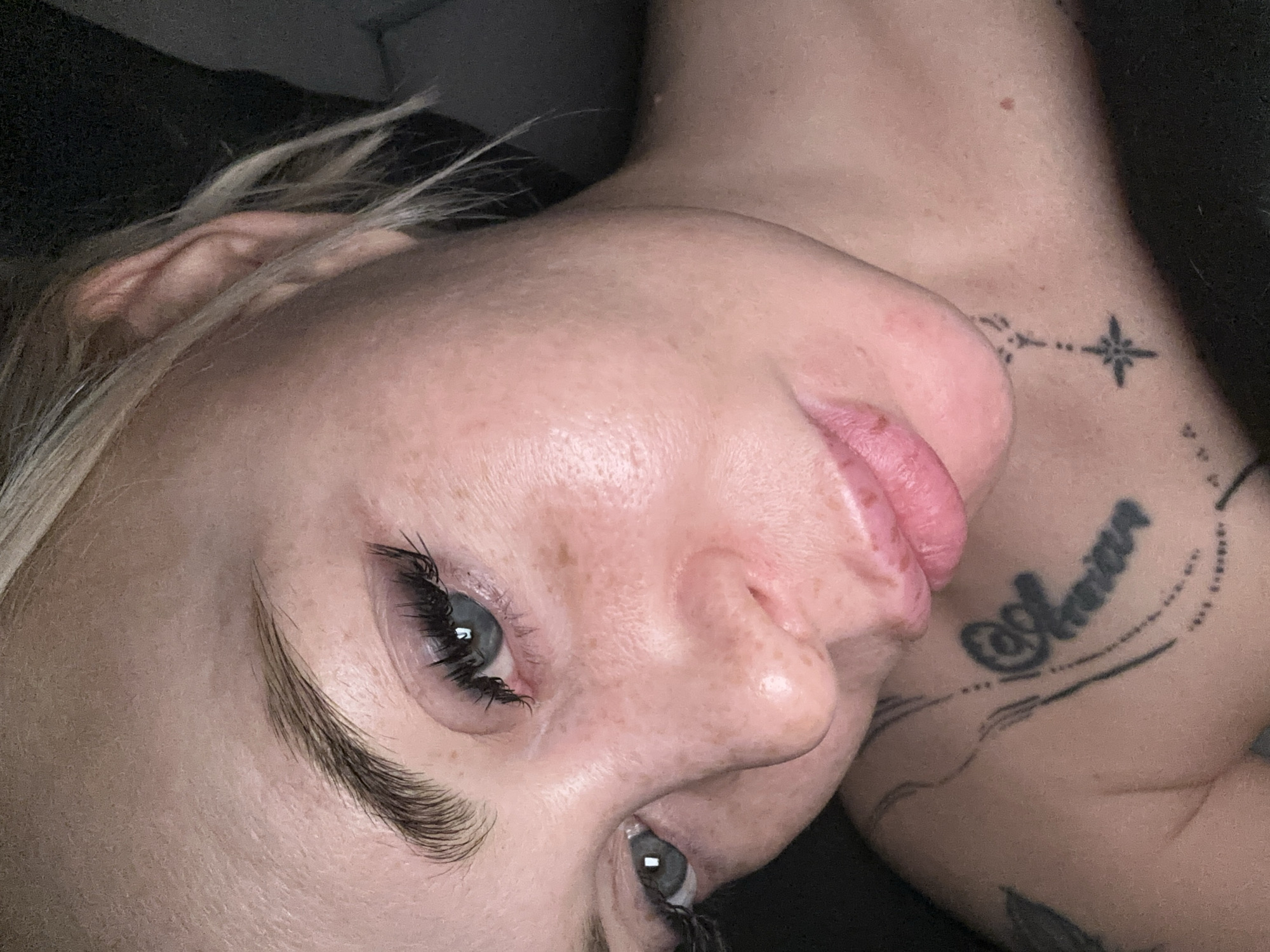 A close-up, slightly overhead, and rotated portrait features a person with fair skin, light blue-grey eyes, and blond hair, lying down in what appears to be an indoor setting with a dark background. The person's head is tilted, showing their face in profile from the nose down, and a full view of one eye, while the other eye is partially visible.
Notable facial features include a scattering of freckles across the nose and cheeks. The visible eye has prominent, dark eyelashes, likely enhanced with mascara or extensions, and a well-groomed, natural-looking eyebrow. The lips are full, pink, and slightly parted, appearing soft and perhaps slightly wet. Light-colored hair strands are visible on the left side of the frame, near the forehead.
On the person's neck and upper shoulder, several tattoos are visible. One design on the neck resembles a small compass rose or starburst pattern, connected by a dotted line. Another, larger tattoo on the shoulder features cursive script. The visible text clearly reads "@Amara". Below this, some faded lines and partial designs suggest other tattoos. The overall lighting is soft, highlighting the skin texture and details of the face and tattoos.