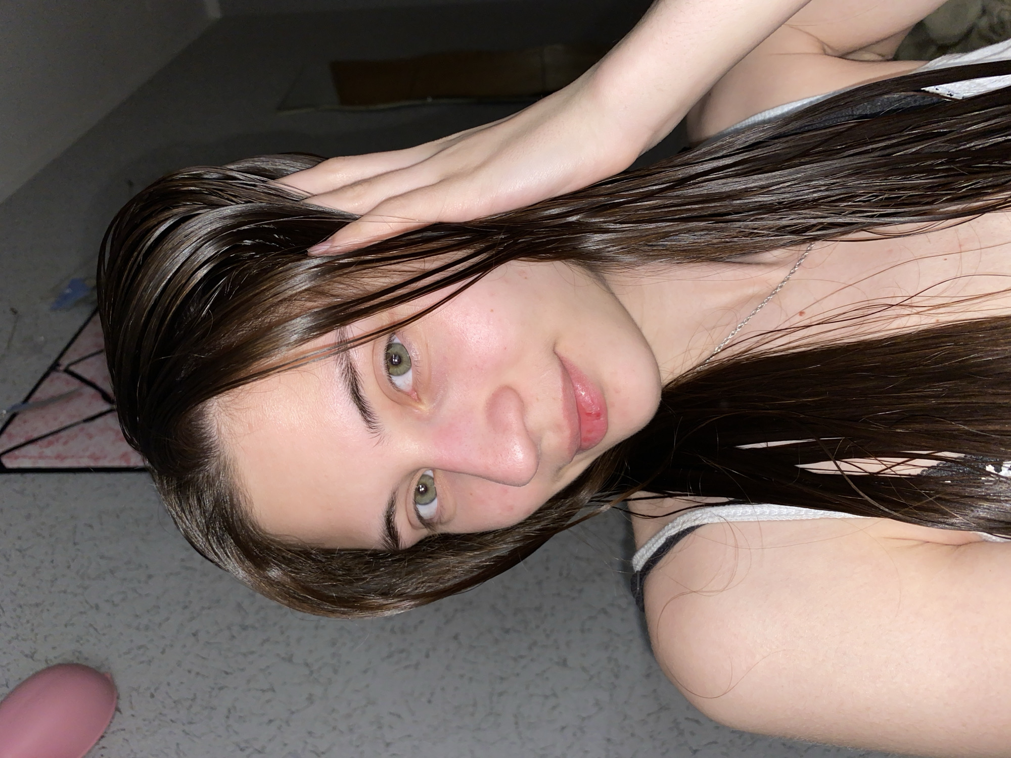A young woman with dark, wet hair is indoors. She is wearing a tank top with a black and white strap, and a silver necklace is visible. Her skin is fair, and she has green eyes. Her hand is placed on her head, moving her hair. The location is inside an unidentified building, possibly in the city of Votkinsk, Russia, although no identifying elements indicate the specific city. The background is a grey carpet and a dark wall, and a pink object.