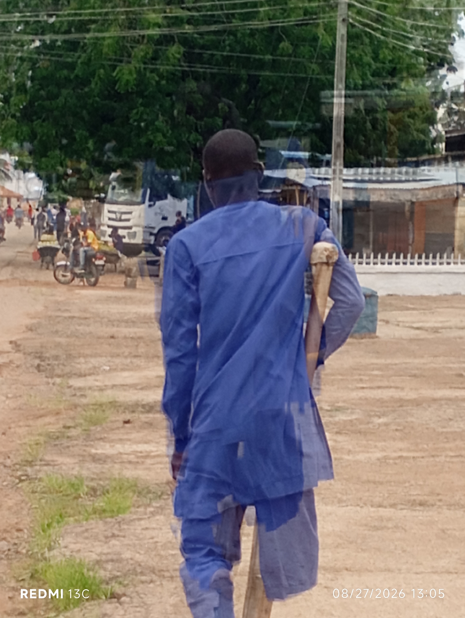 A man wearing a blue robe and pants walks away from the camera in an outdoor, dusty environment. He is using a wooden crutch for support, holding it in his right hand and resting it against his right shoulder. The crutch appears to be made from a branch or root. In the background, there are several buildings, some with corrugated metal roofs, and a large white truck. A few motorcycles and people are visible in the middle ground, suggesting a marketplace or busy street scene. Lush green trees are present further in the background, and utility lines are strung overhead. The overall impression is a slice of everyday life in Shendam, Nigeria.