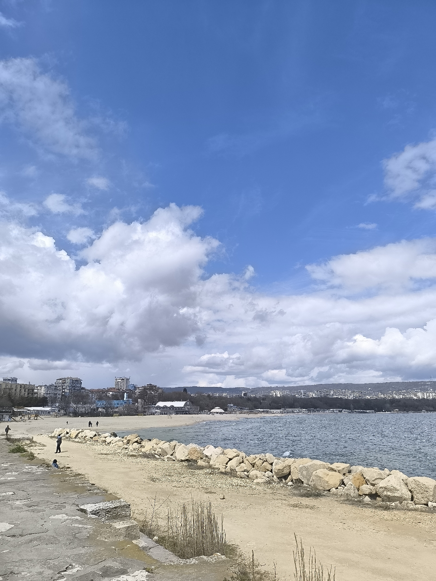 The image depicts a coastal scene in Varna, Bulgaria. A stone-paved pathway leads to a sandy area bordered by a rock barrier separating it from the Black Sea. Several buildings are visible along the shoreline in the background, indicating an urbanized setting. A few people are walking along the beach. The sky is partially covered with large, white and grey clouds. The overall scene appears to be a casual seaside environment with a mix of natural and urban elements.