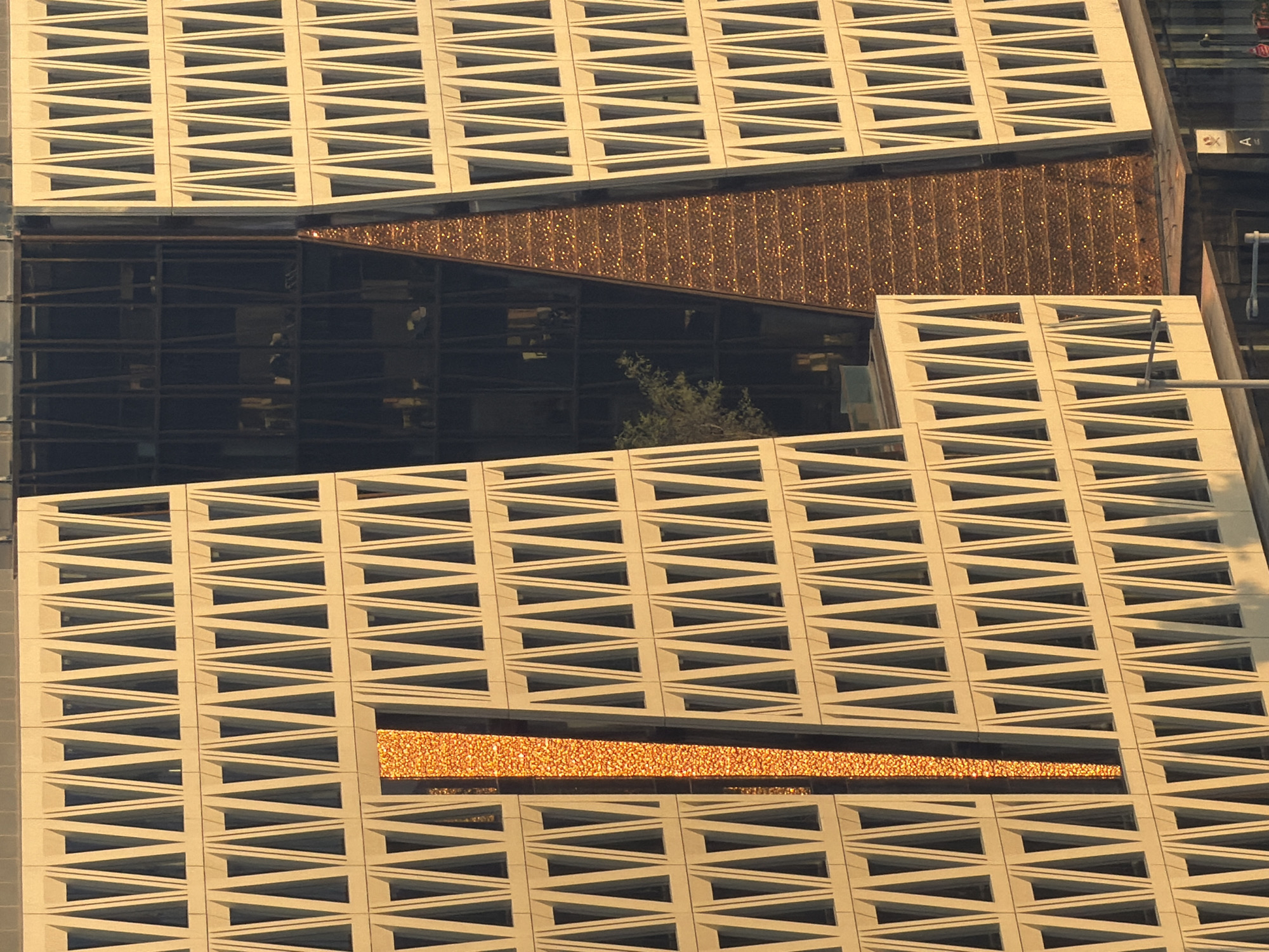The image shows a close-up, eye-level view of a modern building exterior, possibly an office building, in an unknown urban location. The facade is predominantly light-colored, with a repeating pattern of triangular indentations that create a series of narrow, vertical windows. A prominent, vertical section of the building is covered in a textured, shimmering orange material, reflecting the sunlight and appearing granular or glittery. To the right of this section, another part of the building has a dark, glass-paneled facade, revealing interior elements like office spaces and lights. A small, leafy tree is visible through the glass. The overall impression is one of architectural design with contrasting materials and textures.