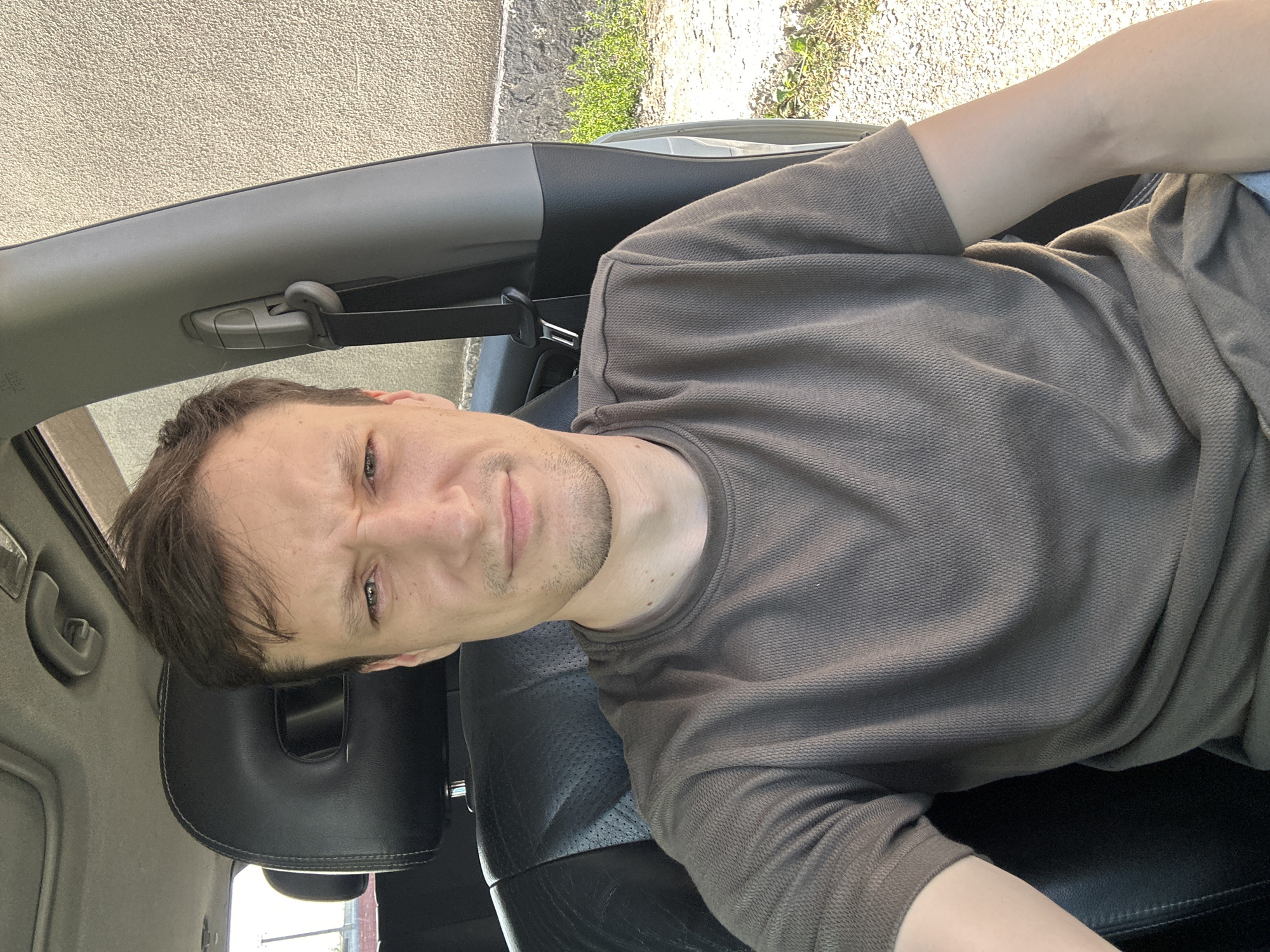A man is sitting in the driver's seat of a car, looking at the camera with a concerned or questioning expression. He is wearing a brown t-shirt. The interior of the car is visible, including the headrest, seat, and part of the door panel with a seatbelt. Outside the car, a textured wall and some greenery are visible. The image appears to have been taken on a sunny day. The specific location of Shatava, Ukraine is not determinable from the visual information in the image.