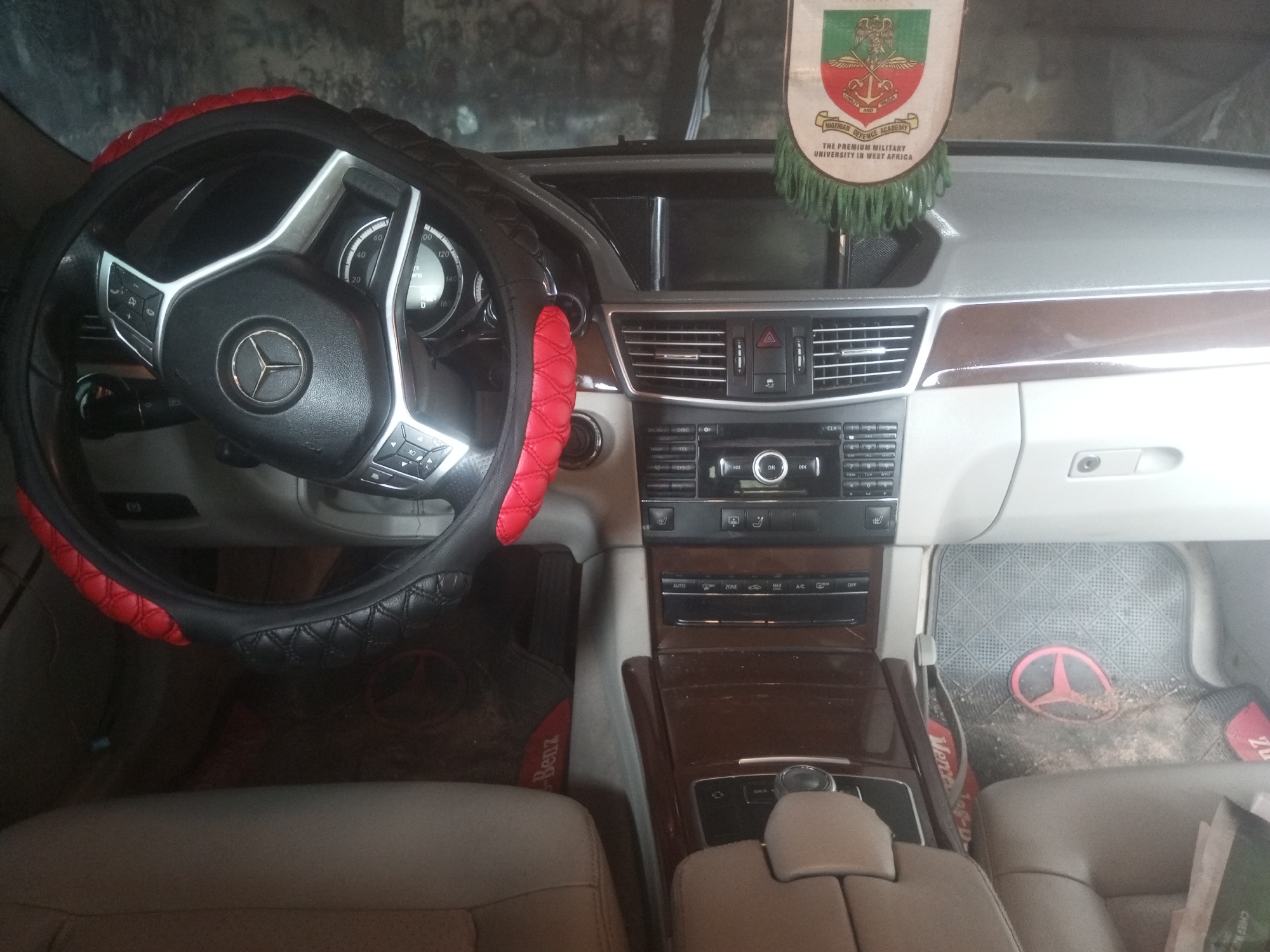 The image shows the interior of a Mercedes-Benz vehicle, likely a E-Class model, in New Stadium, Nigeria. The steering wheel is covered with a black and red quilted steering wheel cover. The dashboard features a central infotainment screen, air vents, and climate control buttons. A Nigerian Military University banner hangs from the rearview mirror. The floor mats are also branded with the Mercedes-Benz logo. The car appears to be parked in a garage or covered area.