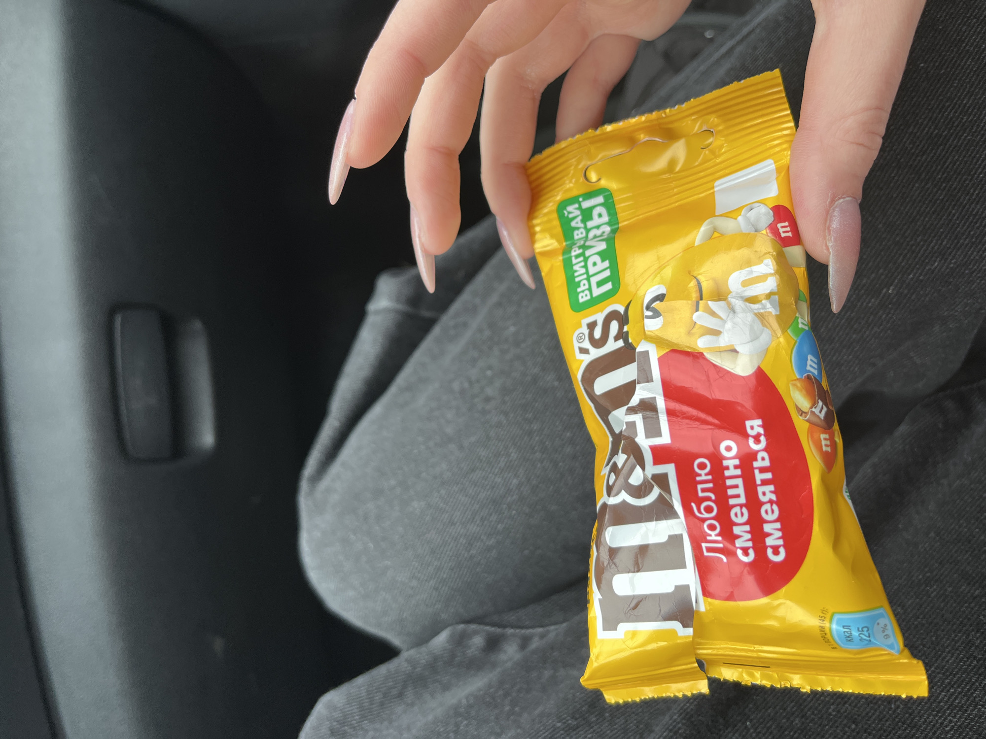 A close-up, eye-level shot shows a person's hand with long, light-pink acrylic nails holding a yellow M&M's candy bar wrapper. The wrapper features the M&M's logo in brown and white, a red oval with Russian text that reads "Люблю смешно смеяться" (I love to laugh funny), and a cartoon M&M character. A green banner on the wrapper says "ВЫИГРЫВАЙ ПРИЗЫ" (Win prizes). The lower left corner of the wrapper displays nutritional information: "В 1 ПОРЦИИ (45 г):" (per serving (45g):), "ККАЛ 225" (kcal 225), and "9%". The background consists of dark grey denim pants and a dark grey car interior. The image is cropped to focus on the candy and hand, with no discernible city or Russia context present within the frame.