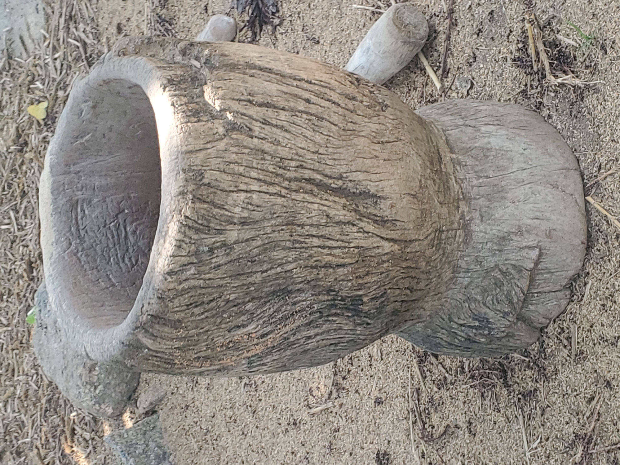 A close-up shot shows a weathered, hollowed-out wooden mortar resting on sandy ground. The mortar's exterior is rough and textured with visible grain patterns and vertical striations, indicating age and use. The opening at the top is circular and appears to be worn smooth. The surrounding environment consists of dry, light-colored sand mixed with small twigs and debris. In the background, a larger, rough-textured stone or wooden object is partially visible to the left of the mortar. Two cylindrical wooden pieces lie on the ground to the right and behind the mortar. The lighting suggests it is daytime, with a bright highlight visible inside the mortar. The scene is located in Dawaki, Nigeria.