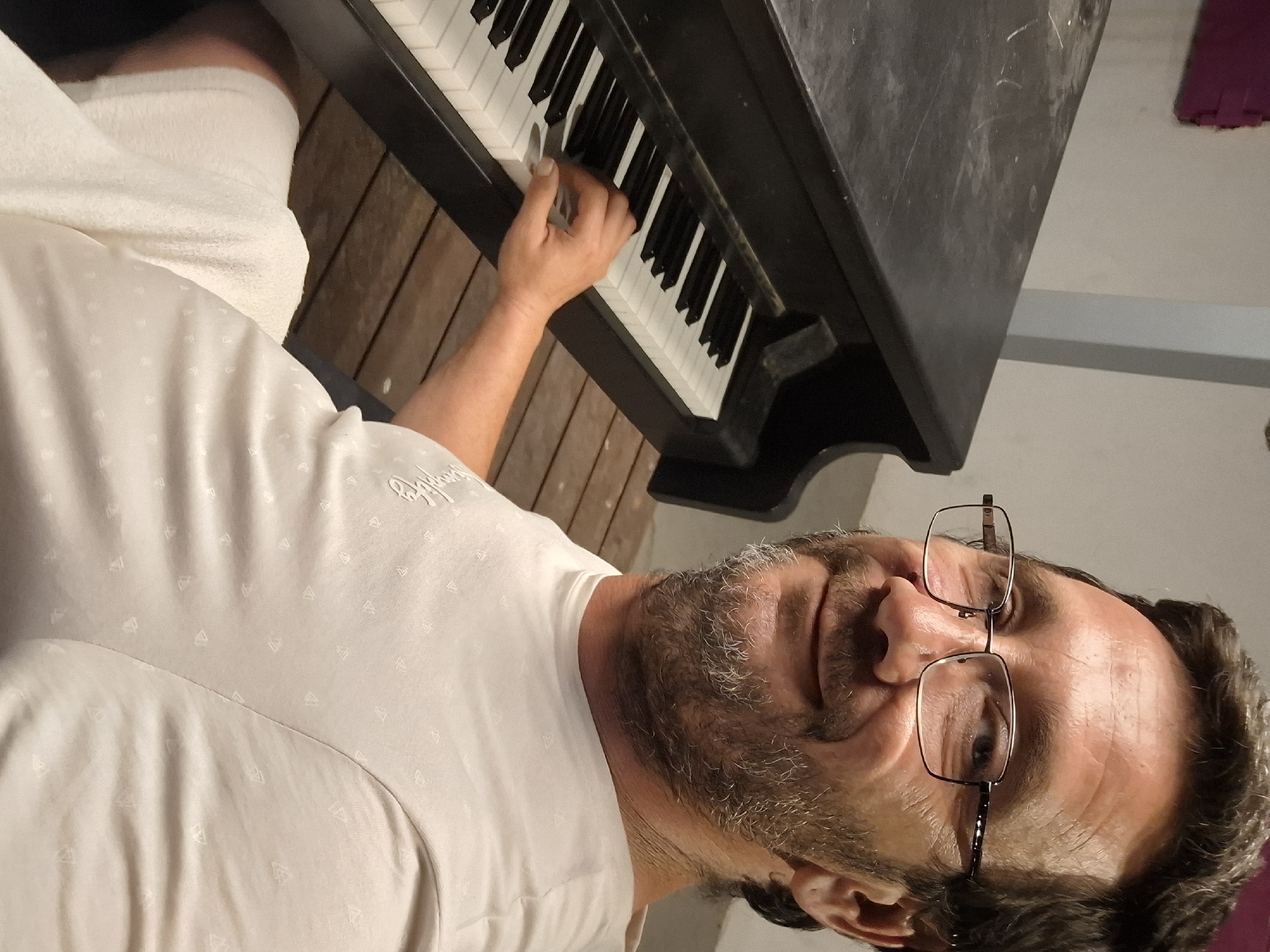 A man with glasses and a beard is sitting at a piano, with his right hand on the keys. He is wearing a light-colored t-shirt and white shorts. The piano is black and appears to be old and dusty. The background shows a wall and wooden floor. The image was taken in Yerocham, Israel, likely in a residential setting.