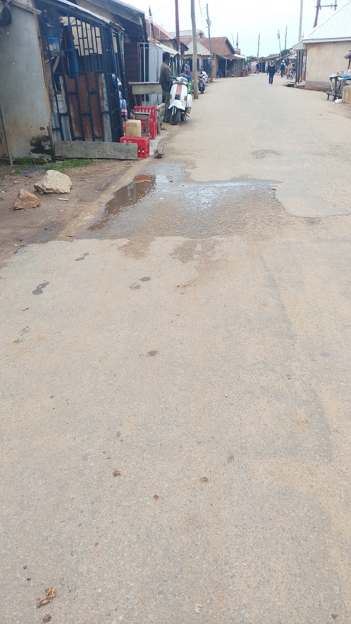 The image depicts a street scene in a place that appears to be a developing area, possibly in a rural or semi-urban setting. In the foreground, a wet patch of road reflects the overcast sky. The road surface is asphalt and shows signs of wear and tear, with some scattered debris. Footprints are visible in the wet area and on the drier parts of the road.
To the left of the road, there are several structures that appear to be market stalls or small shops. These structures have corrugated metal roofing and some have open fronts with metal grates. In front of one of these structures, there are several red plastic crates, some empty and some with items inside. A white scooter is parked next to the crates, and a person is standing behind it, facing away from the camera.
Further down the street, more similar structures line both sides. People can be seen walking and standing near these establishments. Another scooter is parked further down the street. The overall environment suggests a bustling, though perhaps not highly developed, commercial area. The sky is overcast, indicating potential for rain or recent rainfall.