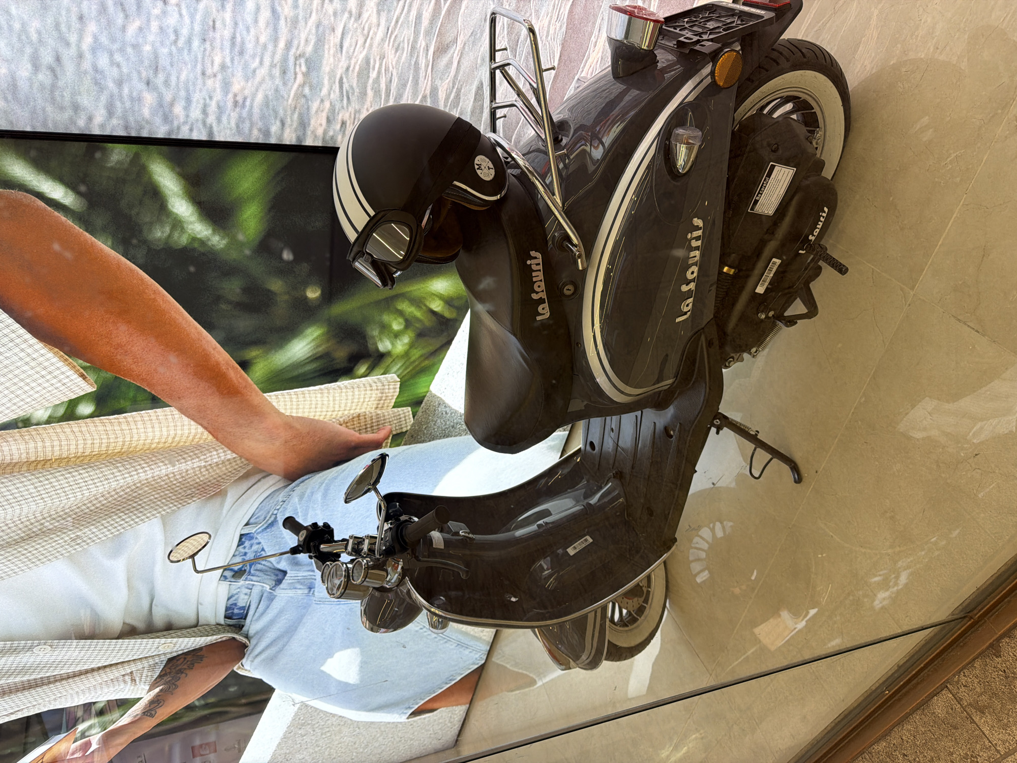 A close-up shot shows a La Souris scooter on display, possibly in a shop window in Amsterdam, Netherlands. The scooter is dark gray with chrome accents and white-walled tires. A retro-style helmet rests on the scooter. The scooter is positioned in front of a large printed backdrop featuring an image of a person wearing light-colored clothing. The background suggests a bright, possibly outdoor scene with greenery. The foreground appears to be a reflective surface, such as a glass window or shiny floor.