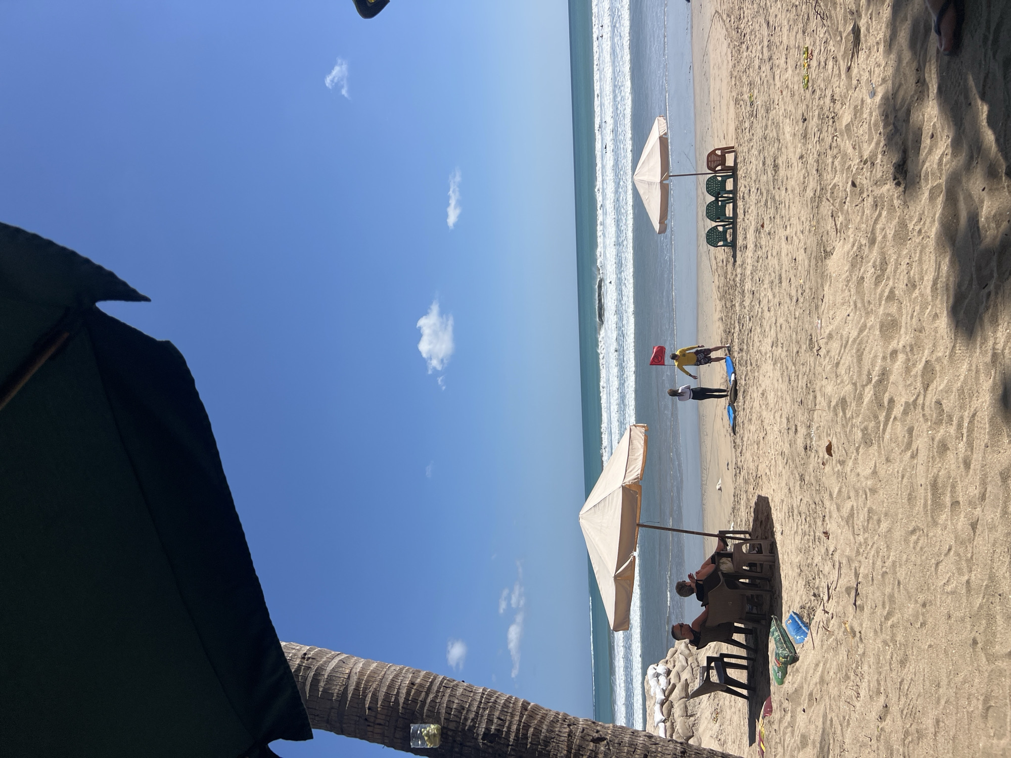 A bright, sunny day is depicted at Kuta Beach, Bali, Indonesia, with the ocean stretching out under a clear blue sky dotted with a few scattered clouds. In the foreground, a large dark green umbrella partially obscures the upper left corner of the frame, casting a shadow that extends across the sandy beach. To the left, the rough trunk of a palm tree stands tall, with a small glass jar containing what appears to be a drink with ice and a straw attached to it.
On the sand, two people are seated in chairs under a light-colored umbrella, facing the ocean. The chairs are dark brown plastic, and behind them, several large sandbags are piled up. Further back on the beach, a second light-colored umbrella is set up, with three green plastic chairs and one orange plastic chair placed in front of it.
Two individuals are standing near the water's edge. One, wearing a yellow shirt and patterned shorts, is holding a red flag on a pole. Another person, dressed in a white top and dark leggings, stands next to them. Several surfboards are visible on the sand near them.
The ocean features moderate waves breaking, with numerous surfers scattered across the water. The shoreline is marked by wet sand near the water, transitioning to drier, rippled sand further up the beach. Small pieces of debris are scattered across the sand in the middle ground. The overall scene conveys a relaxed beach atmosphere.