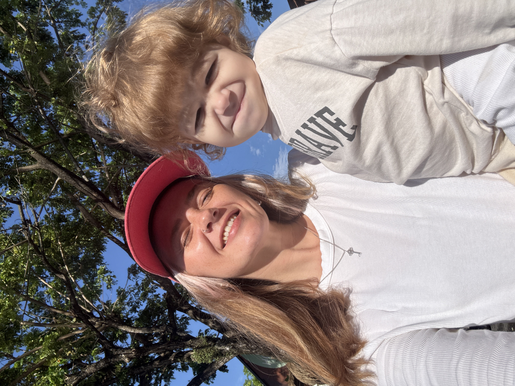 In a close-up, sun-drenched selfie taken outdoors, a woman with blonde hair and a pink baseball cap smiles with her eyes closed. She wears a white t-shirt with a silver key pendant necklace. Beside her, a young child with curly blonde hair makes a funny face, wearing a light-colored long-sleeved shirt with the word "BRAVE" printed on it. The background features lush green trees and a bright blue sky, suggesting a location in Choeng Thale, Thailand, likely during daylight hours.