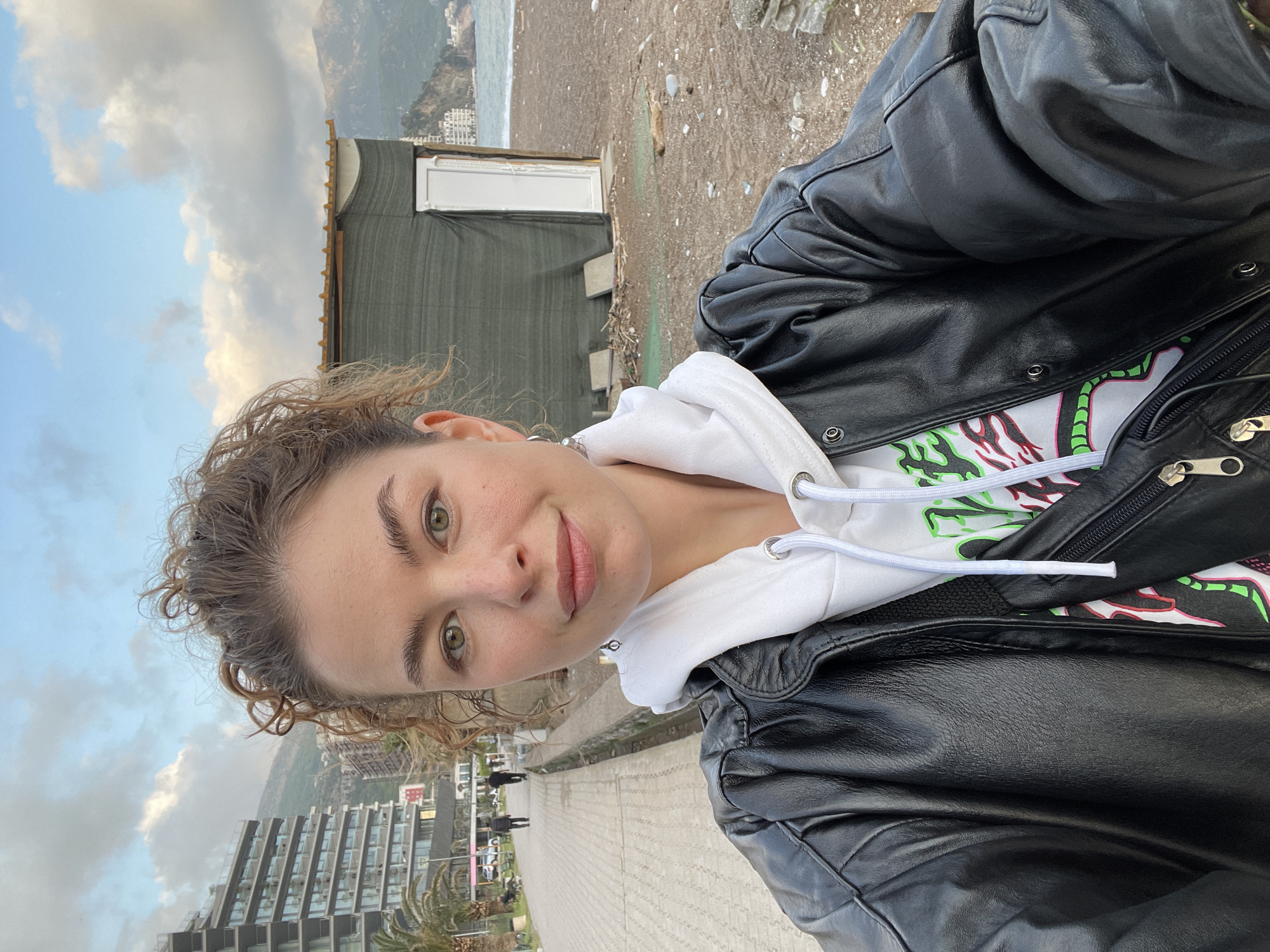 A selfie taken outdoors, showing a fair-skinned person with light brown curly hair, wearing a white hoodie and black leather jacket. The background reveals a coastal setting, likely in the city of Boreti, Montenegro, with a beach, ocean, and distant buildings. A weathered structure with a green facade and a white door is visible near the person. The foreground shows a paved walkway with a few pedestrians in the distance. The sky is cloudy with patches of blue.