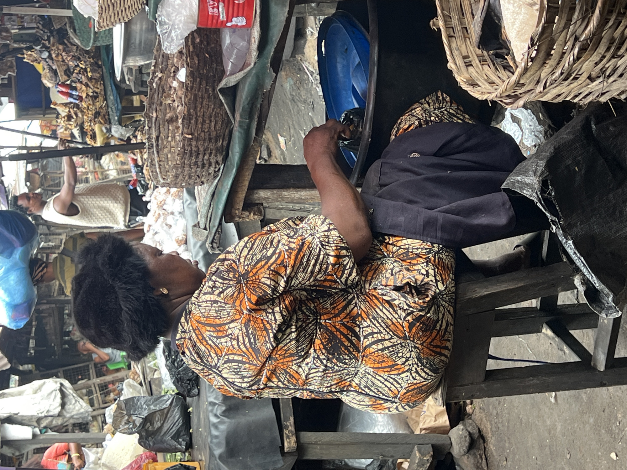 The image shows an individual, likely a woman, seated in what appears to be a bustling market area. She is wearing a patterned orange and black top and is positioned on a dark wooden stool. Her hair is black, and she is wearing earrings. In front of her is a blue basin, and she seems to be engaged in some activity within it.
The setting is crowded with various items typical of a market: baskets, plastic bags, and other unidentified goods. Another person can be seen in the background wearing a white tank top. The lighting is ambient, suggesting an outdoor environment. The image appears to be taken in a street market setting.