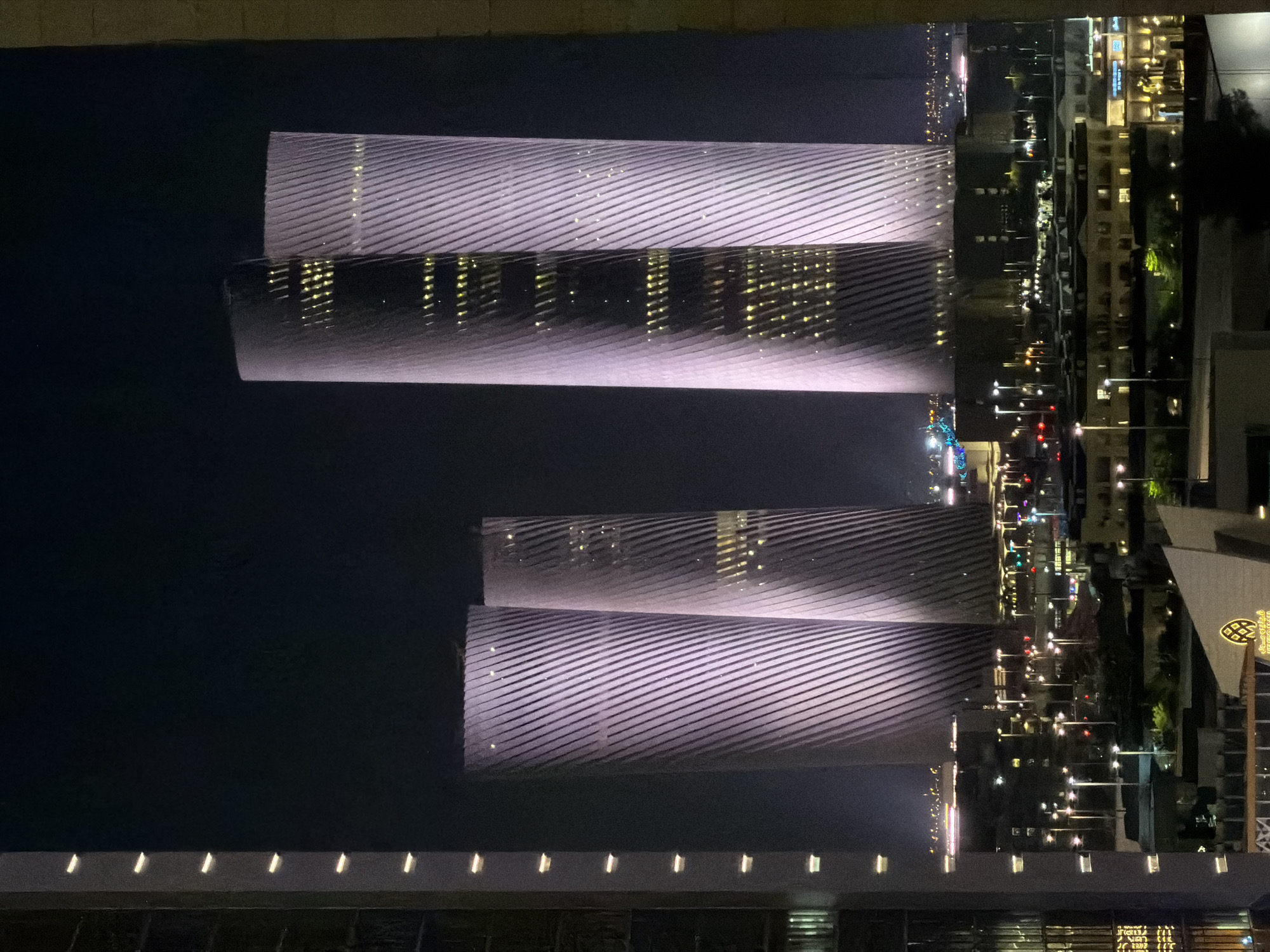The image shows a nighttime view of several modern skyscrapers with distinctive spiraling facades, illuminated in purple and white light. The buildings have a textured appearance due to the diagonal lines of their exteriors. Interior lights are visible through windows, appearing as scattered yellow dots. In the foreground and midground, there are lower-rise buildings and a cityscape with streetlights and various illuminated structures. The sky is dark and clear. The overall impression is an urban landscape at night, dominated by the imposing presence of the illuminated skyscrapers.
