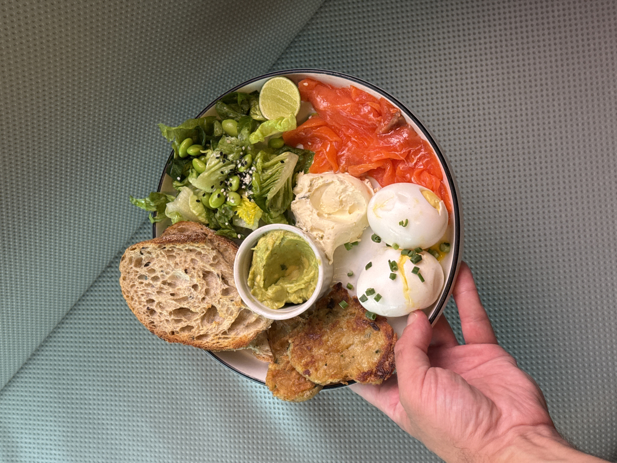 A close-up, overhead shot captures a hand holding a round plate showcasing a diverse breakfast or brunch meal, set against a textured, light teal background. The plate is thoughtfully arranged with various fresh components.
On the upper left, a vibrant green salad of romaine lettuce and edamame is visible, topped with black and white sesame seeds, accompanied by a fresh lime wedge. Adjacent to the salad, several slices of rich orange smoked salmon are neatly placed. In the center of the plate, there's a substantial scoop of a creamy white spread, likely cream cheese.
Below the spread, two perfectly poached eggs, garnished with chopped green chives, are presented, with a hint of golden yolk peeking through one. A small white ramekin holds a portion of mashed avocado or guacamole. Completing the savory items are two golden-brown, pan-fried patties, possibly hash browns or vegetable fritters. On the lower left, two thick slices of rustic, whole-grain bread, appearing to be sourdough, are included.
A person's hand, with a light skin tone, holds the rim of the plate, suggesting presentation or serving. The meal's composition and freshness imply a healthy and gourmet offering, consistent with cafe fare, possibly from North Kuta, Indonesia. There is no visible text in the image.