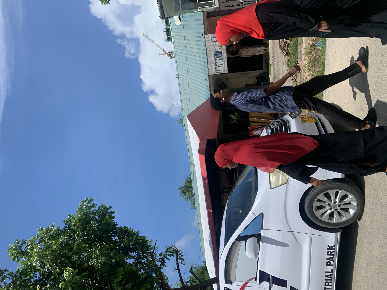 The image depicts a bright, sunny street scene in Dar es-Salaam, Tanzania. The sky is a vibrant blue with scattered white clouds, suggesting clear weather conditions during the daytime.
In the foreground, a white van is partially visible. On its side, the text "TRIAL PARK" is clearly legible, along with a partial logo that appears to be part of "INDUSTRIAL PARK." A woman, dressed in a black abaya and a red hijab, stands near the van, with one hand resting on its front tire.
In the middle ground, a man wearing a blue and white striped shirt and dark trousers is walking or standing on the sidewalk. He has white earbuds in his ears. Behind him, another woman, also in a black abaya and a red hijab, is standing closer to a building. A yellow license plate is visible on a vehicle in the background, possibly belonging to the van or another car.
The background features several buildings, some with distinctive green roofs, consistent with local architecture. Faint text like "WELCOME TO" and "YAMA SAUNA" is partially visible on one of these buildings. A tall yellow construction crane is prominently positioned against the sky, indicating ongoing development in the area. Lush green foliage from trees is visible on the left side of the frame, adding natural elements to the urban setting. The overall scene captures a typical, active street environment.