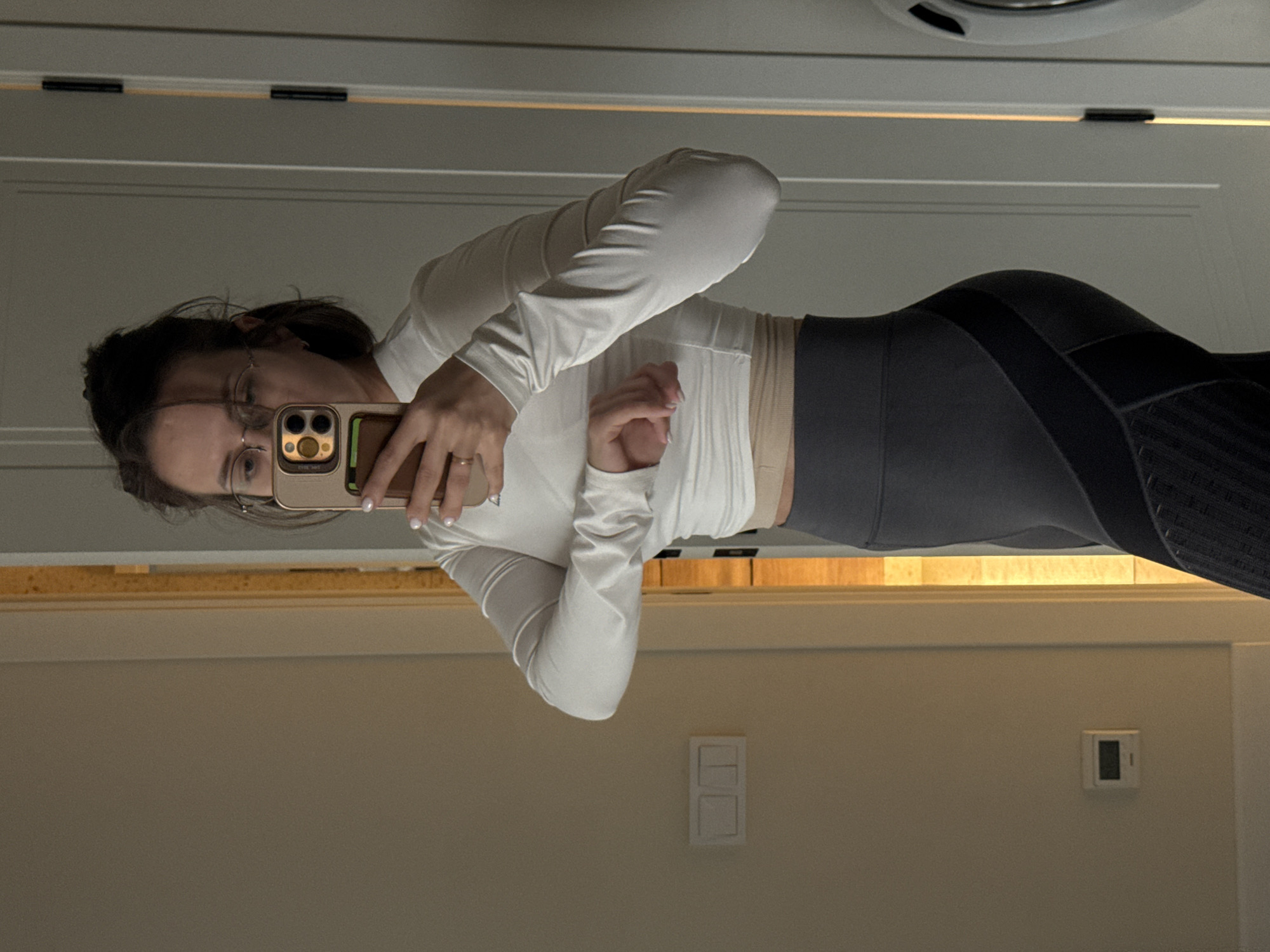 The image is an indoor mirror selfie, capturing a person (likely female) from the waist up. The individual, wearing glasses, is holding a smartphone with a gold-toned case and a multi-lens camera system to capture their reflection. A ring is visible on their left hand.
They are dressed in a white, long-sleeved top and a light, possibly beige, pleated or ribbed camisole underneath. The lower body shows dark grey or black form-fitting leggings with textured or patterned panels on the sides, indicative of athletic wear.
The setting is an indoor room, possibly a hallway or bedroom, with light-colored walls and white trim, all reflected in the mirror. A double light switch or electrical outlet is visible on the wall, along with a smaller rectangular digital display, potentially a thermostat. A light fixture is partially visible on the ceiling above the mirror. The lighting is artificial. The activity is the act of taking a self-portrait via a mirror. No discernible text is visible on objects within the image. This scene takes place in Pervomaysky, Russia.