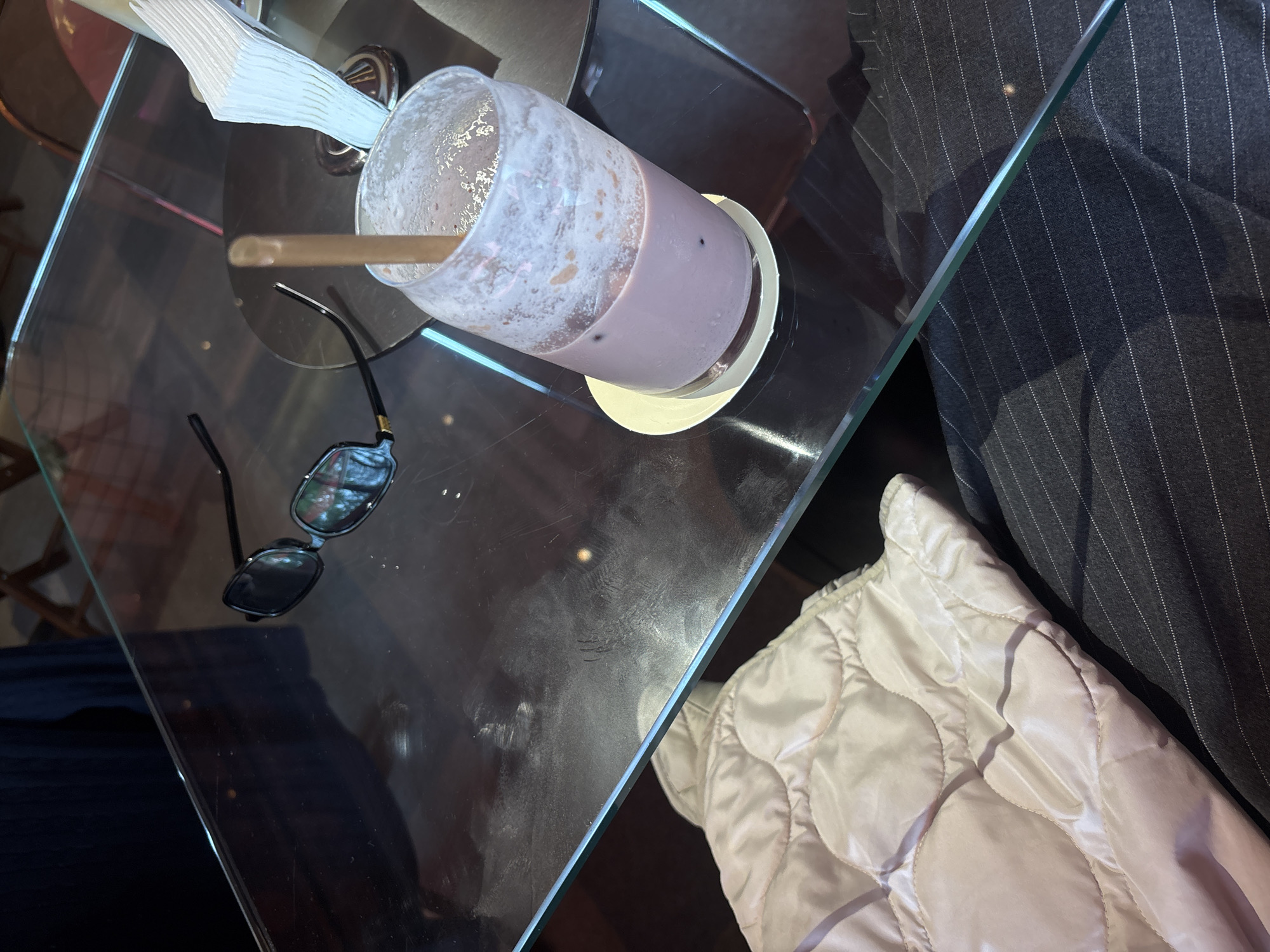 This high-angle shot captures a casual scene on a glass table in what appears to be an indoor setting, likely a cafe or restaurant in Yerevan, Armenia. The central object is a tall glass, mostly empty, containing the foamy remnants of a light purple beverage, possibly a milkshake or smoothie, with dark specks visible. A light brown straw extends from the drink, which rests on a light-colored coaster.
To the left of the drink, a pair of folded black sunglasses with dark lenses is placed on the table. Further back, a stack of white, ribbed napkins or paper serviettes is visible. The clear glass tabletop is highly reflective, showing blurred light sources and indistinct shapes from the surroundings.
On the right side of the frame, a segment of a light-colored, possibly off-white or pale pink, quilted jacket or garment is visible, suggesting a person is seated at the table. Adjacent to the jacket is a dark gray or black fabric with fine white pinstripes, likely another piece of clothing. The background beyond the table is dark and out of focus. The scene suggests a relaxed moment, perhaps after or during a drink. No specific time of day, weather, or emotions are discernible, and no text is visible in the image.