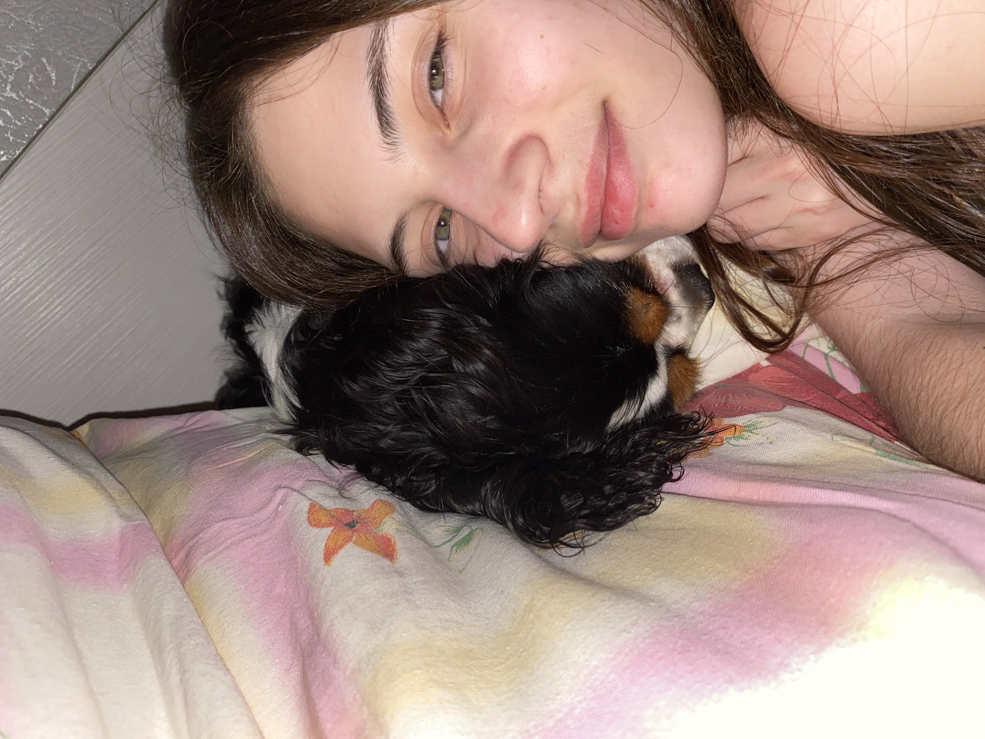 An intimate, close-up indoor shot depicts a young female and a small dog resting together on a bed, likely in Votkinsk, Russia. The female, with long dark brown hair and green eyes, lies on her side, looking towards the camera with a soft, relaxed smile. Her head is nestled against the tri-color dog, which features black, white, and tan fur, and appears to be a Cavalier King Charles Spaniel or similar breed. The dog is mostly obscured by the woman's head and hair but its black, wavy fur and a hint of its face are visible.
The scene conveys a sense of comfort and affection between the human and the animal. The lighting is soft and warm, suggesting it could be evening or night. They are lying on a light-colored duvet or blanket with a tie-dye-like pattern of pinks, yellows, and oranges, and an orange floral design. Part of a plain, light-colored headboard or wall is visible in the top left corner, providing a simple background to the cozy scene. No text is visible in the image.