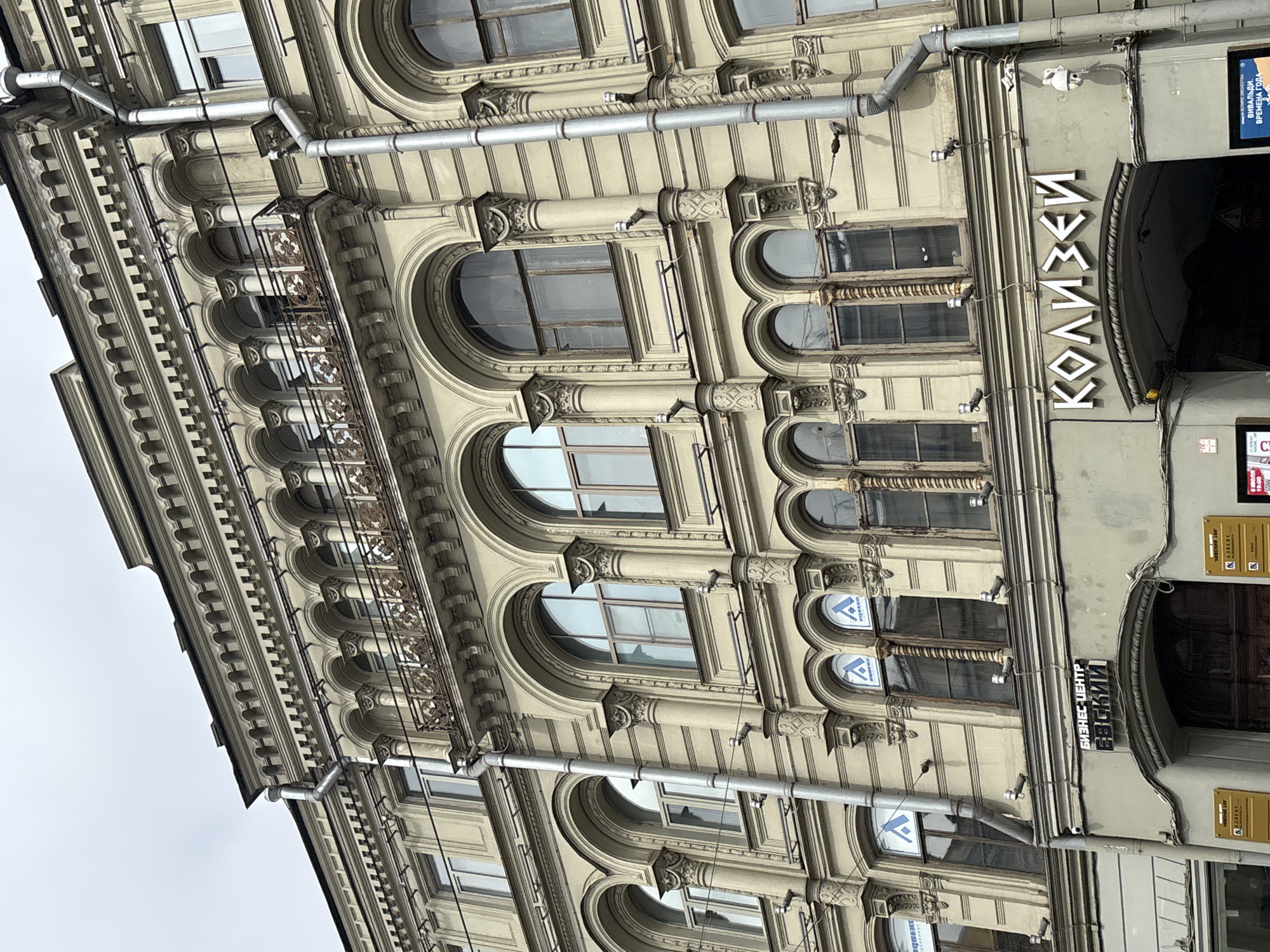 The image shows the facade of a building in Saint Petersburg, Russia, with a prominent sign reading "КОЛИЗЕЙ" (Coliseum) above an arched entrance. The building is characterized by its ornate architecture, featuring multiple levels of arched windows, decorative columns, and a wrought-iron balcony. Rain gutters run vertically along the facade. Several security cameras are visible on the building's exterior. The sky is overcast.